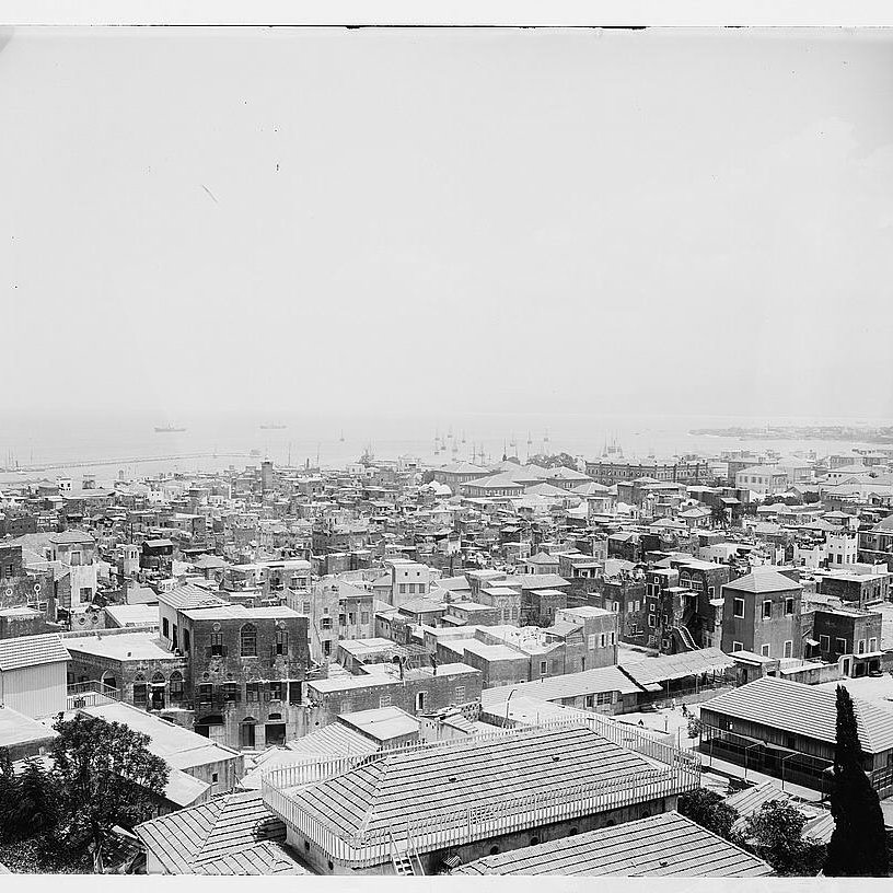 L'image présente une vue panoramique d'une ville côtière. On peut voir de nombreux bâtiments aux toits en tôle, ainsi que des structures en maçonnerie. La mer est visible à l'arrière-plan, avec quelques bateaux amarrés. Les rues semblent étroites et bien dessinées, et il y a des arbres éparpillés dans la zone. L'ensemble donne une impression de densité urbaine et de vie maritime. La photographie semble ancienne, capturant l'architecture et l'ambiance d'une époque révolue.