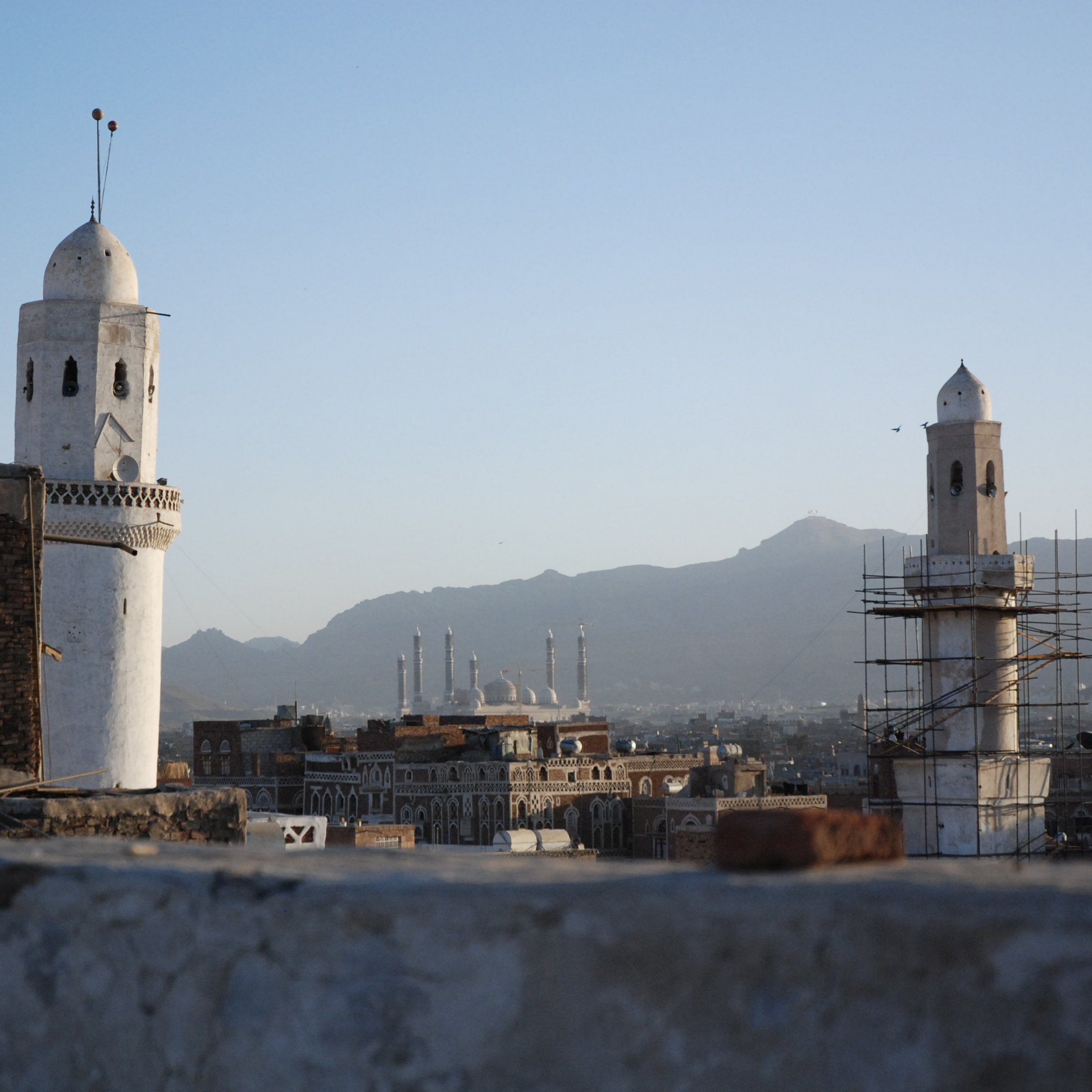 L'image montre une vue panoramique d'une ville avec des minarets distinctifs. On peut voir deux tours blanches caractéristiques, l'une étant en cours de restauration, entourées d'un paysage montagneux en arrière-plan. Le ciel est clair, indiquant une ambiance paisible au crépuscule. Au loin, plusieurs autres structures architecturales, probablement des mosquées, se dessinent à l'horizon, contribuant à l'atmosphère culturelle et historique de la scène.