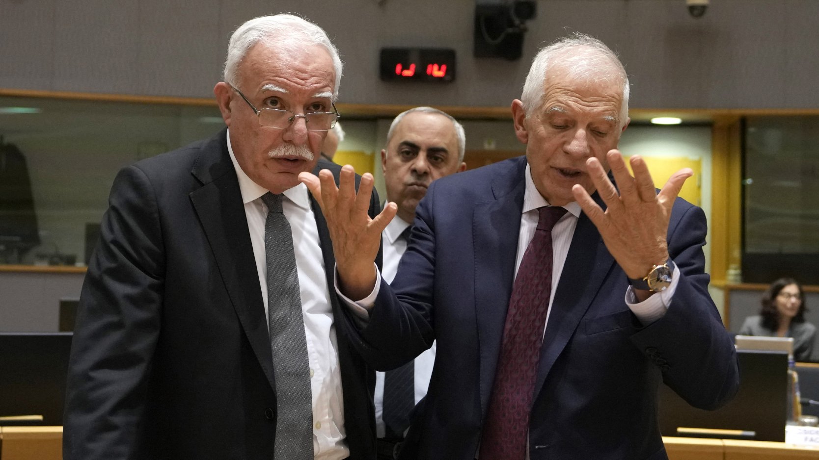 En la imagen se pueden ver a dos hombres en un entorno de reunión, posiblemente en una conferencia o un encuentro diplomático. Ambos parecen estar en una conversación intensa, con gestos de las manos que indican expresión o énfasis en lo que están discutiendo. Al fondo, hay otra persona observando. El ambiente es formal, con un espacio de trabajo y asientos visibles.