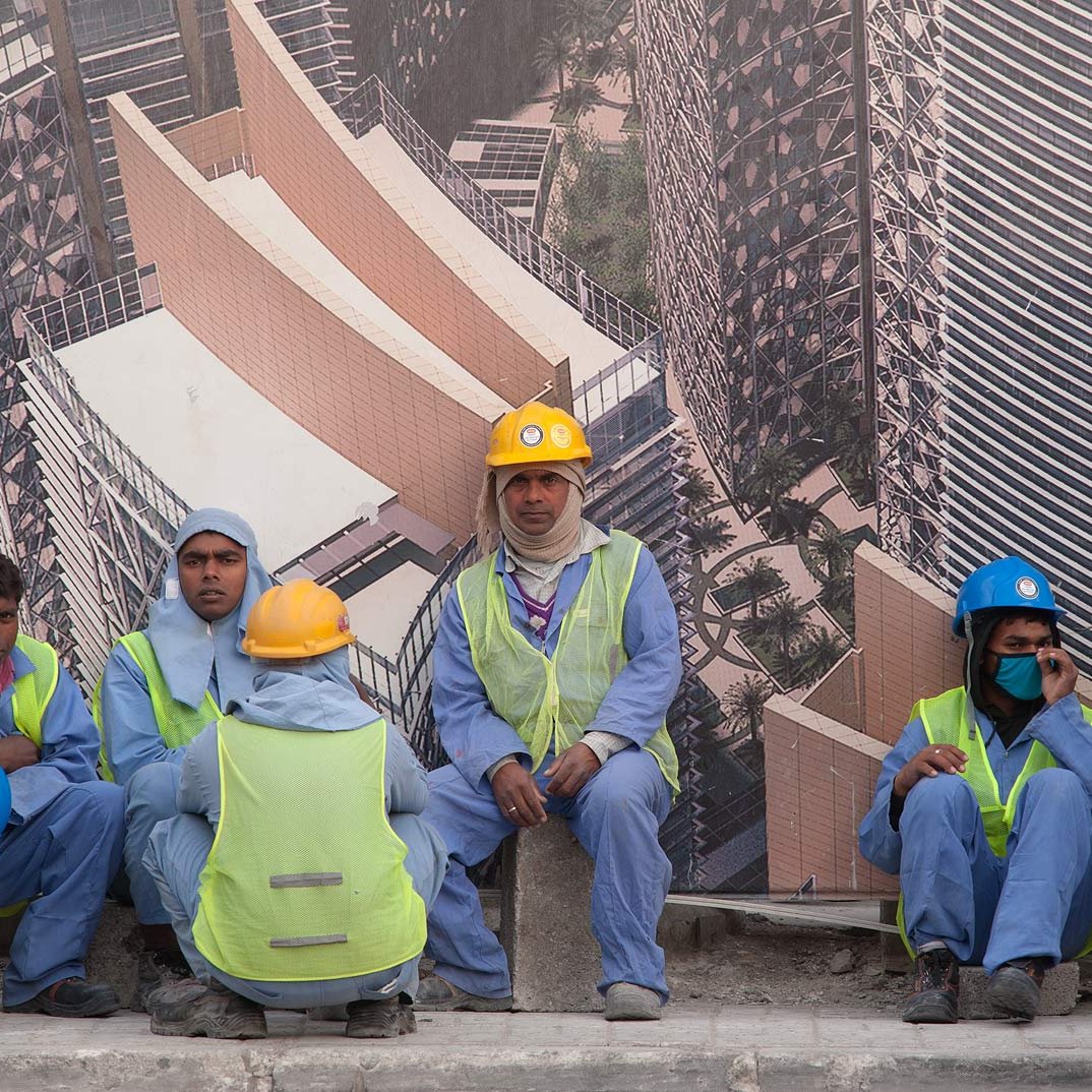 L'image montre un groupe de travailleurs assis sur un mur, probablement en train de faire une pause. Ils portent des vêtements de travail, notamment des gilets de sécurité et des casques de différentes couleurs. L'arrière-plan présente une grande affiche ou un panneau représentant un bâtiment moderne, suggérant qu'ils travaillent sur un site de construction. L'environnement semble urbain et dynamique.