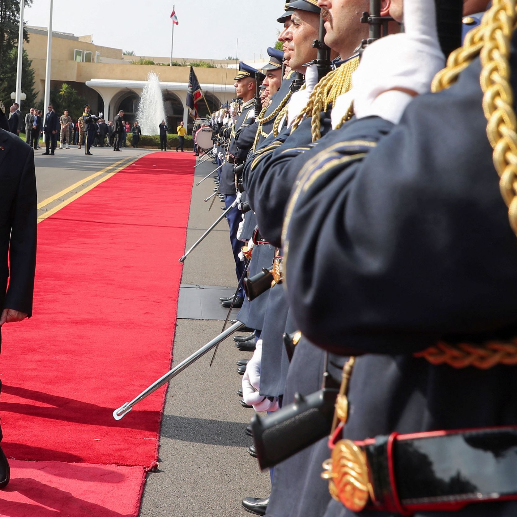 در این تصویر، یک مرد مسن با کت و شلوار تیره در حال راه رفتن بر روی فرش قرمز است. دو طرف او، گروهی از سربازان در لباس‌های رسمی و با تفنگ‌های خود ایستاده‌اند و به او احترام می‌گذارند. در پس‌زمینه، یک ساختمان رسمی و عناصر معماری نمایان است. به نظر می‌رسد که این مراسم یک استقبال رسمی باشد.
