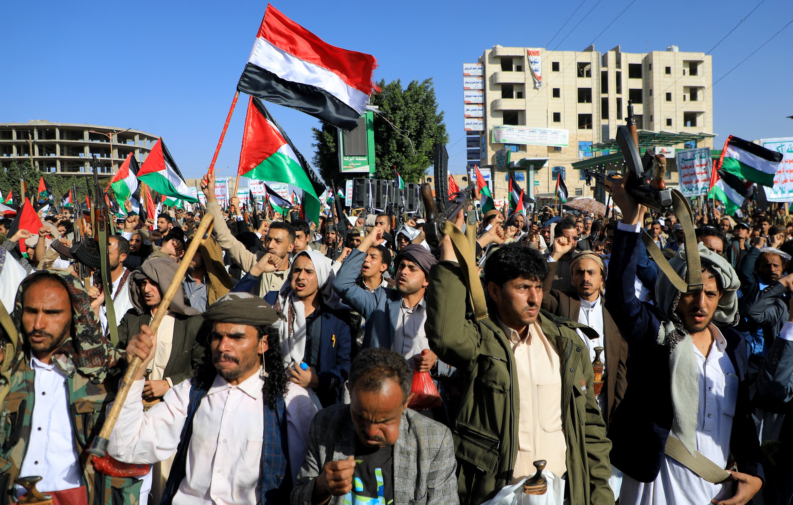 L'image montre une grande manifestation, probablement organisée par une foule, avec de nombreuses personnes levant des drapeaux, notamment des drapeaux aux couleurs de la Palestine. Les manifestants semblent déterminés et engagés, certains brandissant des banderoles. On peut voir des bâtiments en arrière-plan, suggérant un environnement urbain. L'atmosphère semble chargée d'énergie et de passion pour la cause.