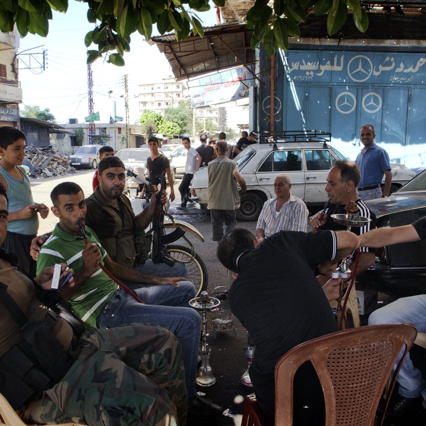 L'image montre un groupe d'hommes assis dans une rue animée. Certains d'entre eux portent des vêtements décontractés et semblent discuter ou se détendre ensemble. On peut voir un homme armé avec une arme à feu, ce qui suggère une ambiance tendue ou de conflit. En arrière-plan, il y a des voitures et des bâtiments, ainsi qu'une enseigne avec le logo d'une marque automobile. L'atmosphère semble informelle, malgré la présence d'armes.