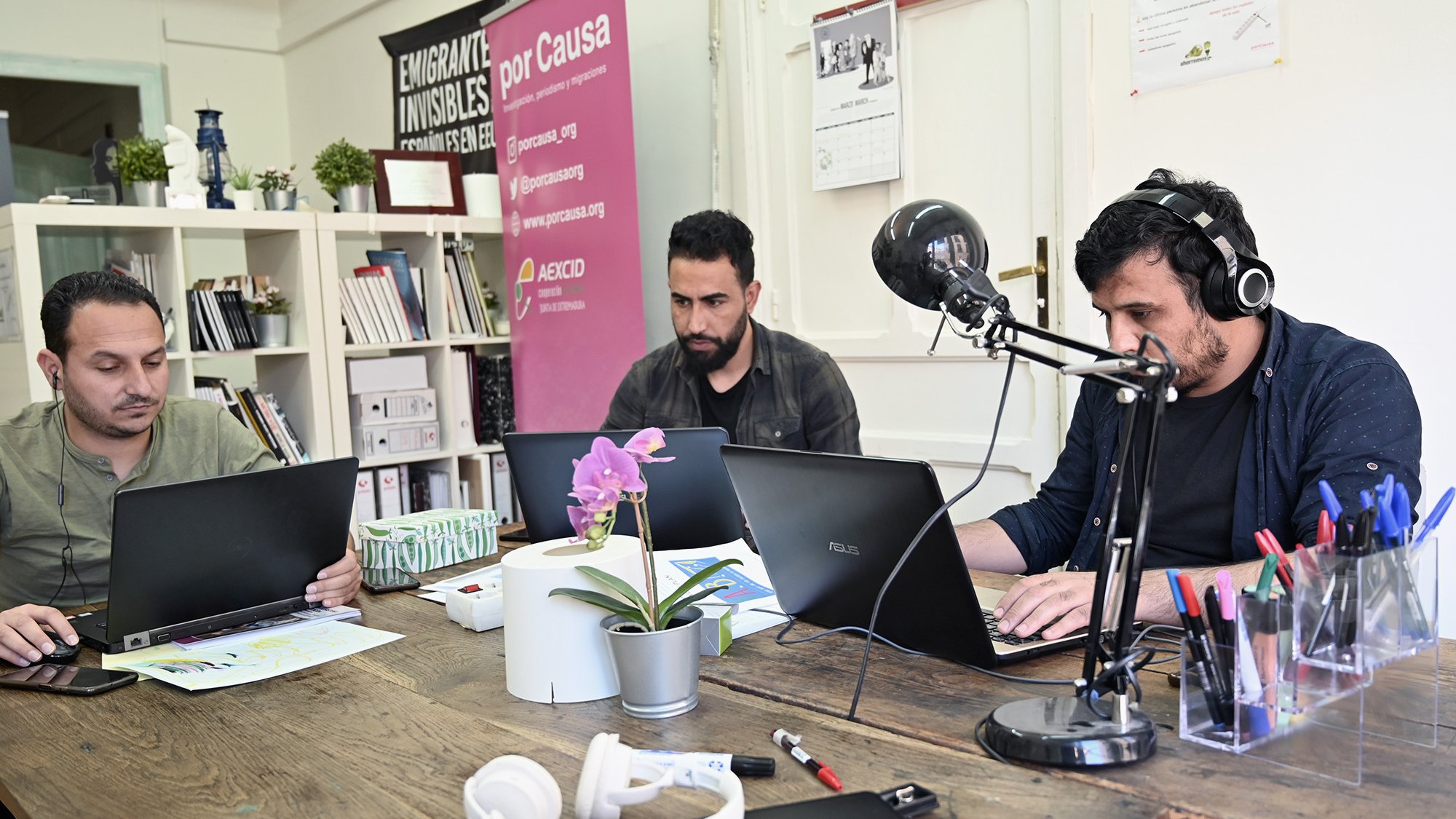 En la imagen se pueden ver a tres hombres sentados en una mesa de trabajo. Cada uno tiene un portátil frente a él y parece que están concentrados en sus tareas. La mesa está decorada con una planta en una maceta y hay auriculares sobre la superficie. En el fondo, hay una pared con algunos carteles, incluyendo uno que dice "Emigrantes Invisibles". También hay estantes con libros y otros materiales. La atmósfera sugiere un ambiente de trabajo colaborativo y dinámico.
