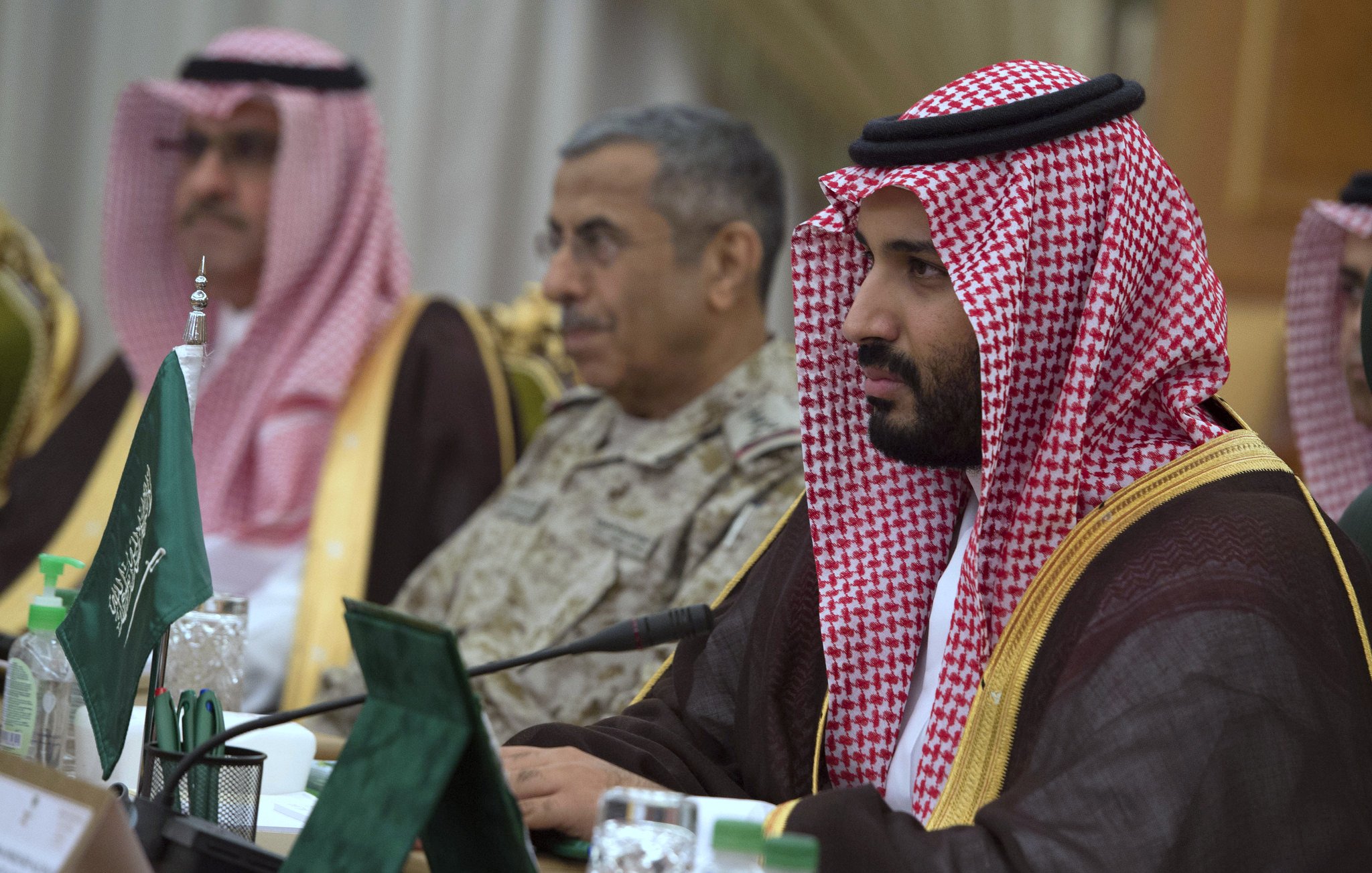 The image shows a group of individuals in formal attire, seated around a table. The man in the foreground is dressed in traditional Saudi attire, wearing a red and white checkered headscarf (ghutrah) and a long brown robe. He appears focused, looking ahead. In the background, another man in military attire is also present, along with other officials. The setting appears to be a formal meeting or conference, indicated by the presence of flags and documents on the table.