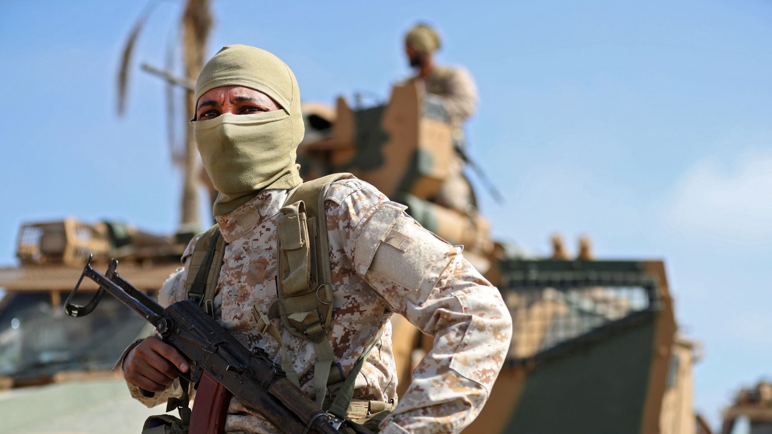 L'image montre un soldat portant un uniforme camouflé et un masque couvrant le bas de son visage. Il est équipé d'une arme et se tient en position vigilante, probablement en mission. En arrière-plan, on peut apercevoir un véhicule militaire et un autre soldat. Le ciel est dégagé, suggérant une ambiance ensoleillée et une atmosphère de tension.