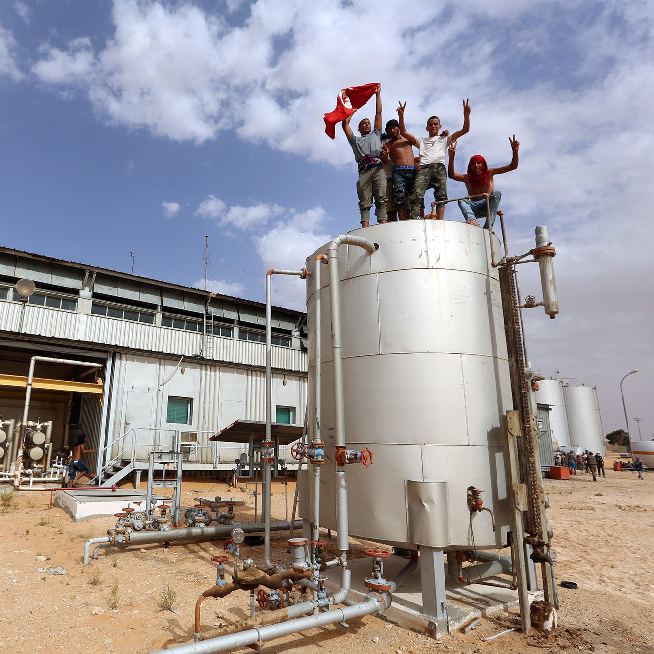 L'image montre un groupe de personnes célébrant en se tenant sur un réservoir, probablement dans une installation industrielle. Ils brandissent un drapeau et affichent des signes de joie. En arrière-plan, on peut voir des infrastructures industrielles et un ciel nuageux. Le sol est désertique, ce qui suggère une ambiance aride. L'ensemble de la scène semble évoquer un moment de fête ou de revendication.