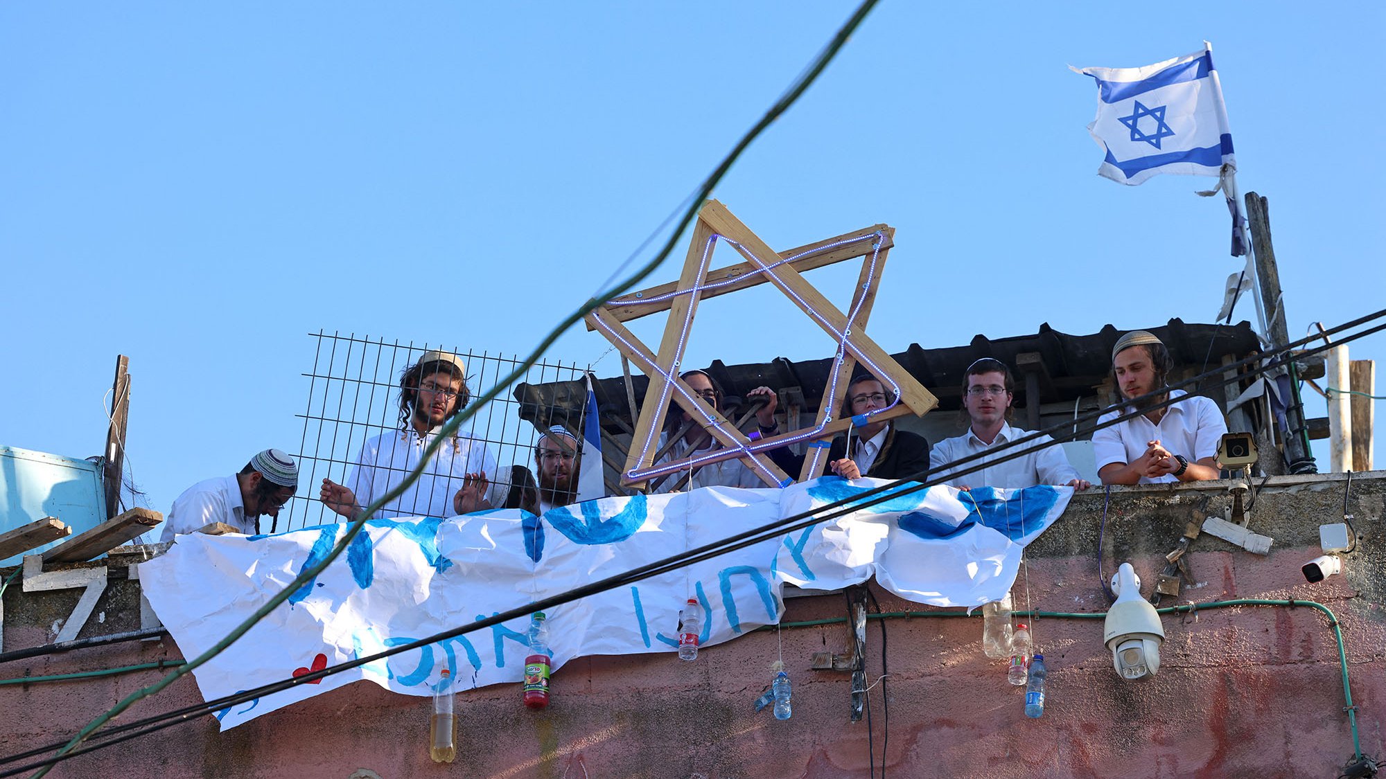 L'image montre un groupe de personnes rassemblées sur un toit. Elles portent des vêtements traditionnels, et certaines tiennent un symbole en forme d'étoile de David. En arrière-plan, il y a un drapeau israélien flottant. On peut également voir une banderole accrochée, suggérant un message ou une revendication. Le ciel est dégagé, et il y a une ambiance de rassemblement communautaire.