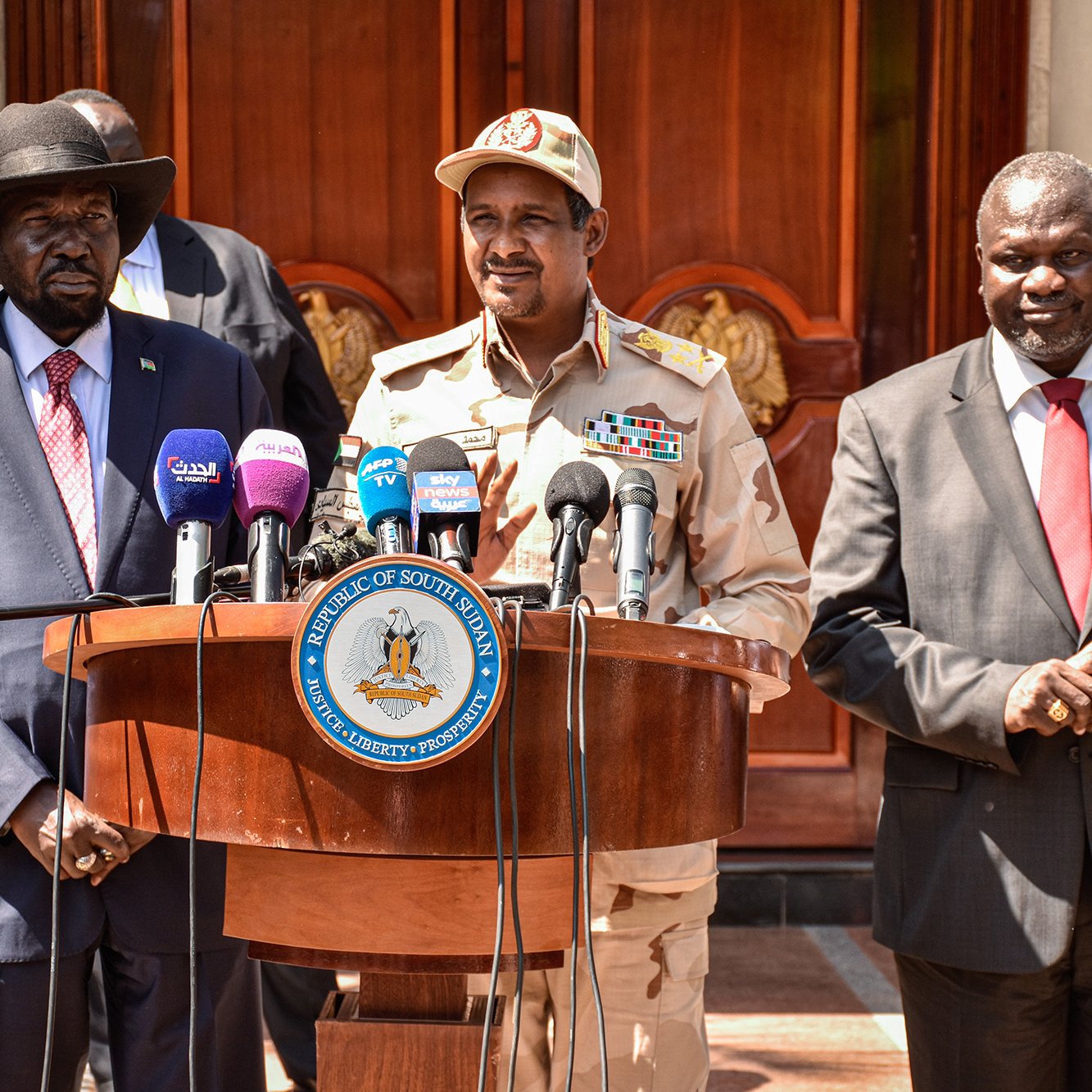 The image shows a group of three men standing behind a wooden podium. The man on the left is wearing a black hat and a suit with a red tie. The man in the center is wearing a military uniform with various insignia and a cap. The man on the right is dressed in a dark suit. They appear to be at an official event, likely addressing the media, with microphones positioned in front of them and a decorative building in the background.