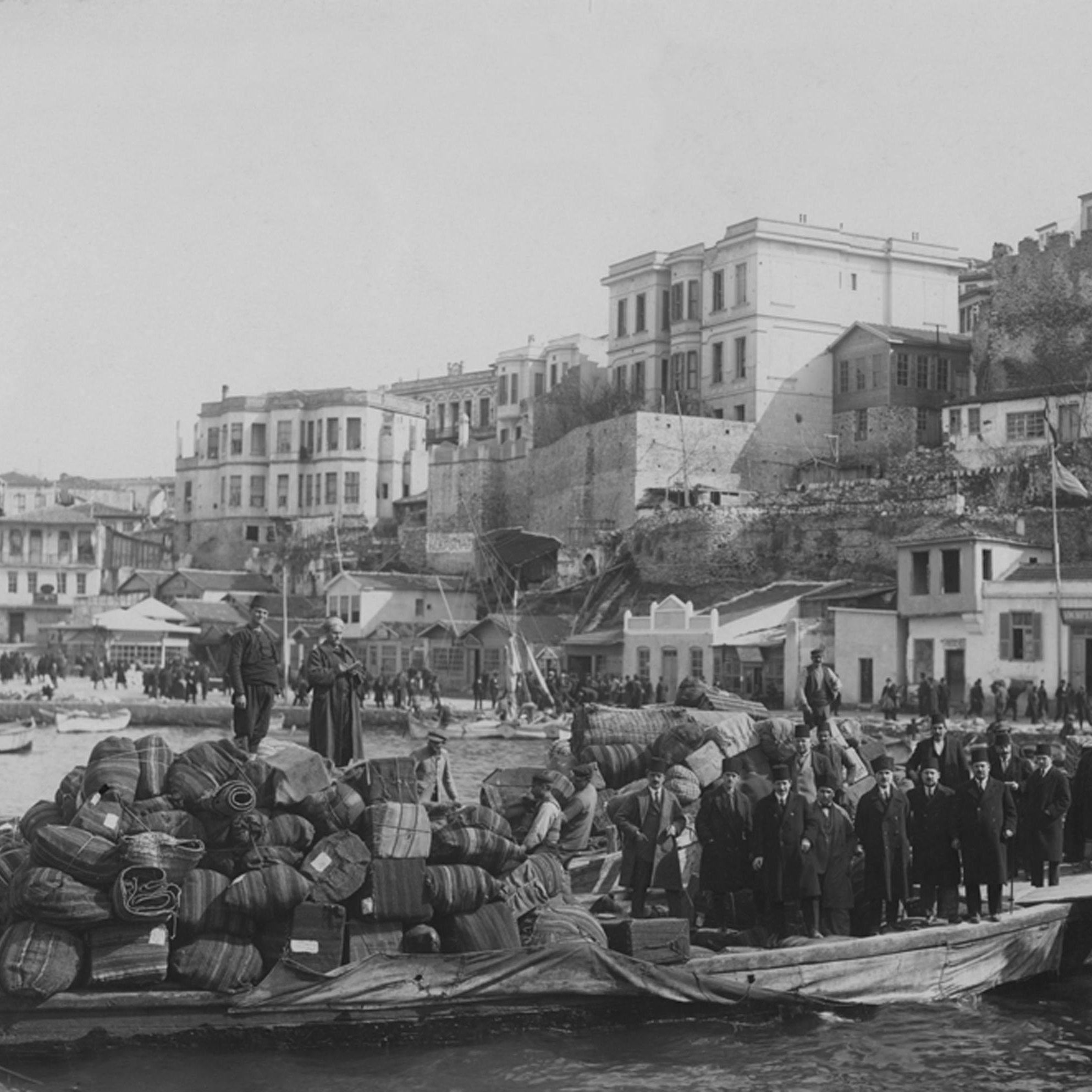 Cette image en noir et blanc montre une scène portuaire historique. On peut voir une barge chargée de marchandises, notamment des fûts et des sacs, avec des personnes debout dessus. En arrière-plan, il y a des bâtiments qui semblent faire partie d'une ville côtière, avec des structures en pierre et des maisons aux balcons. La côte est animée, avec d'autres bateaux et des gens sur le rivage, créant une atmosphère de commerce maritime. La lumière et le contraste évoquent une ambiance d'époque.