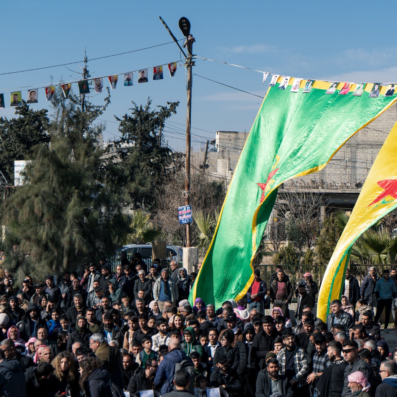 در این تصویر، تعدادی از افراد در یک تجمع یا مراسم حضور دارند. در پس‌زمینه، پرچم‌های بزرگ با رنگ‌های سبز و زرد به همراه ستاره‌ای قرمز به اهتزاز درآمده‌اند. مردم در حال ایستادن و تماشا کردن هستند و درختان و ساختمان‌هایی در دوردست مشاهده می‌شود. همچنین، پرچم‌های کوچک‌تری به نام‌های مختلف در بالای محل تجمع آویزان شده‌اند. آسمان آبی است و جو به نظر شاداب به‌نظر می‌رسد.
