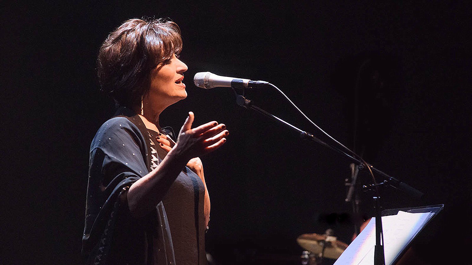 L'image montre une femme sur scène, en train de chanter ou de parler dans un microphone. Elle a les cheveux courts et bruns, et porte une tenue sombre, accompagnée d'un châle. Son expression semble engagée et passionnée, suggérant qu'elle interprète une performance émotionnelle. En arrière-plan, on aperçoit des instruments de musique, ce qui indique qu'elle se produit dans un cadre musical. L'éclairage est tamisé, créant une ambiance intime et captivante.