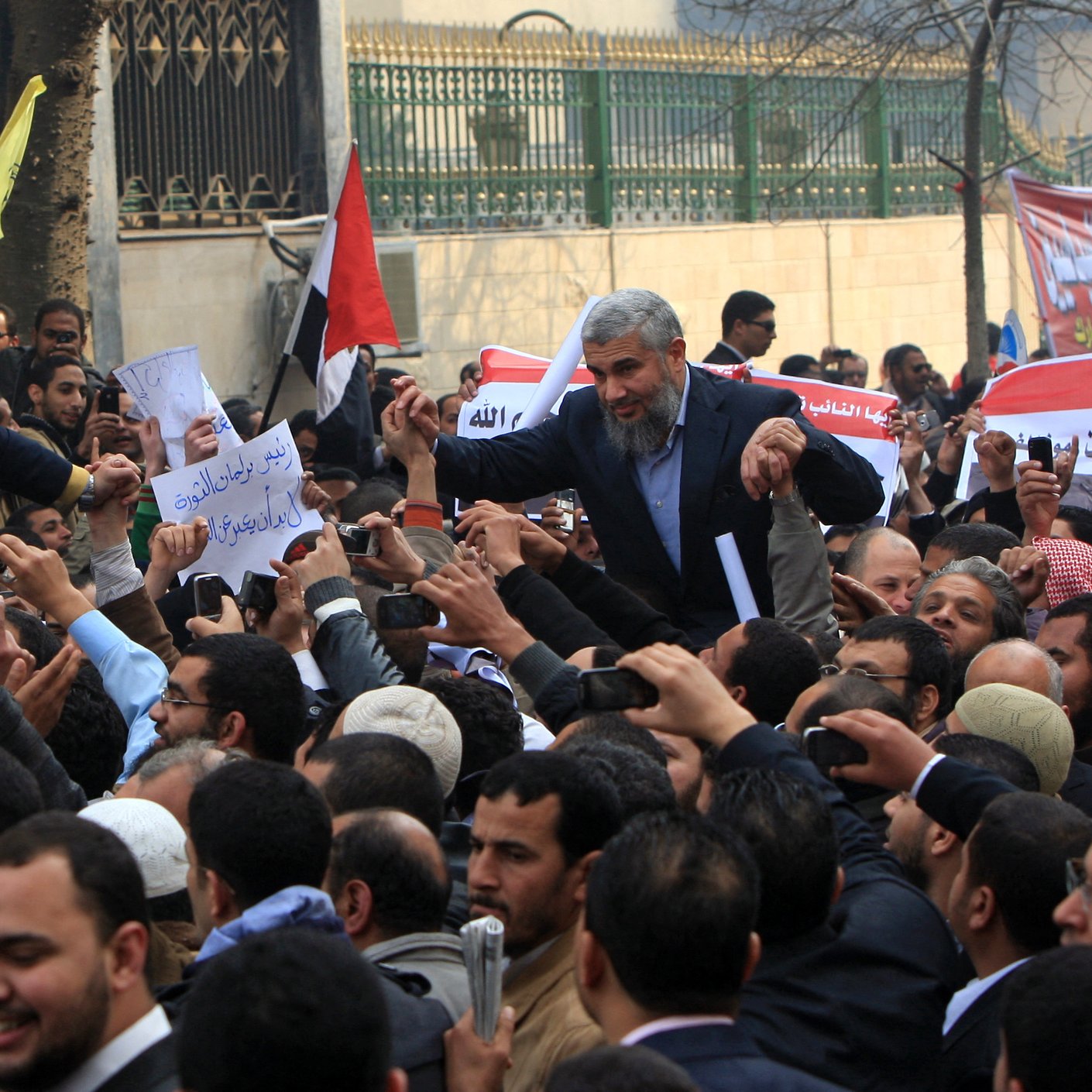 L'image montre une grande foule de personnes rassemblées, probablement lors d'une manifestation ou d'un rassemblement politique. On peut voir des individus tenant des pancartes et des drapeaux, exprimant des opinions ou des demandes. Les expressions des personnes sont variées, allant de l'enthousiasme à la détermination. L'ambiance semble énergique, reflétant un engagement collectif pour une cause. Les drapeaux et les signatures sur les pancartes suggèrent un contexte national ou politique spécifique.