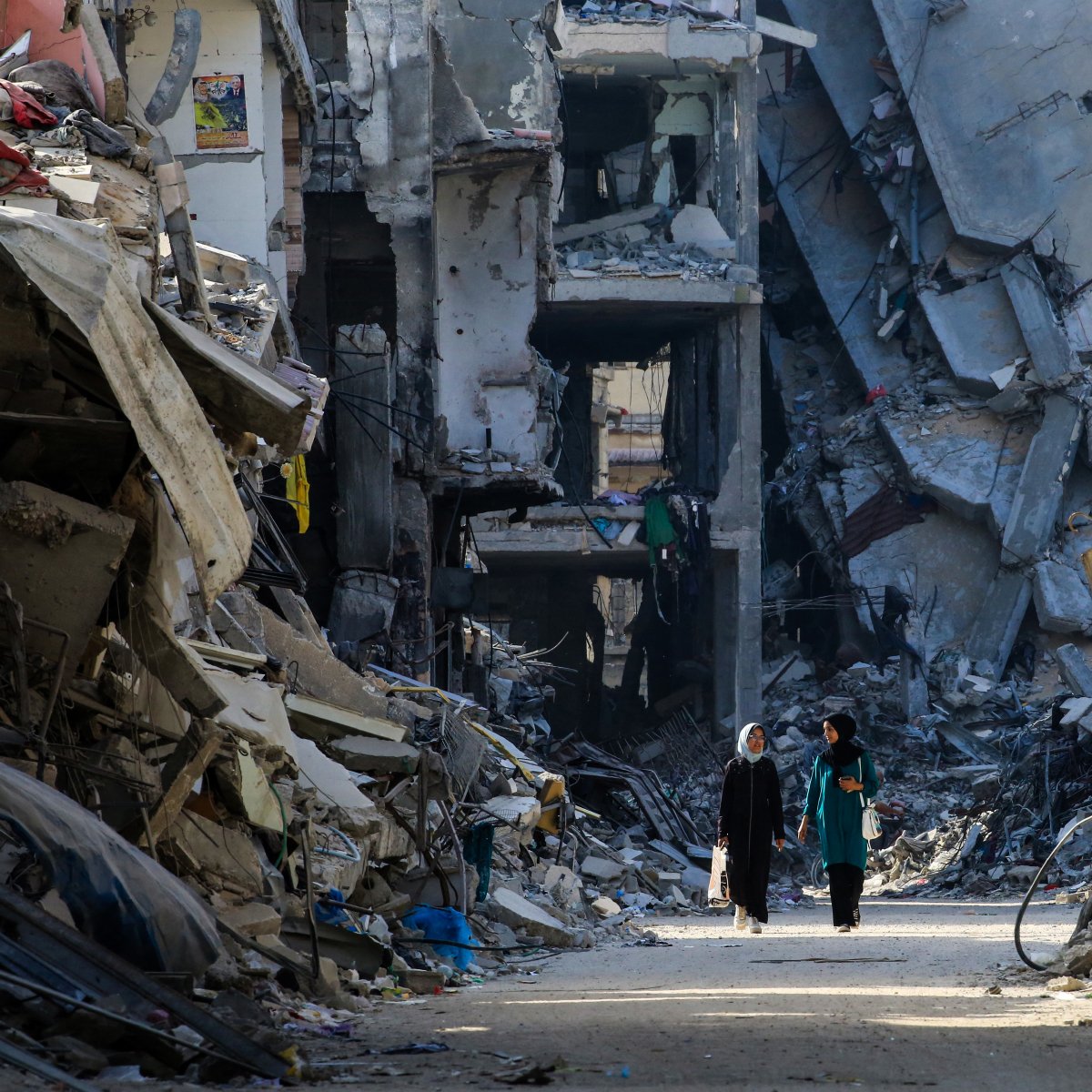 تصویر نشان‌دهنده خیابانی است که در میان ویرانه‌های ساختمان‌ها قرار دارد. دیوارهای بتنی و تخریب‌شده در دو طرف خیابان وجود دارند و debris و زباله‌ها در زمین پخش شده‌اند. دو زن در حال راه رفتن در این خیابان هستند و به نظر می‌رسد در جستجوی امنیت یا کمک هستند. نور خورشید به آرامی از میان ویرانه‌ها عبور می‌کند و سایه‌های طولانی‌ای بر زمین می‌افکند. حس و حال تصویر، غم و ویرانی را به نمایش می‌گذارد.