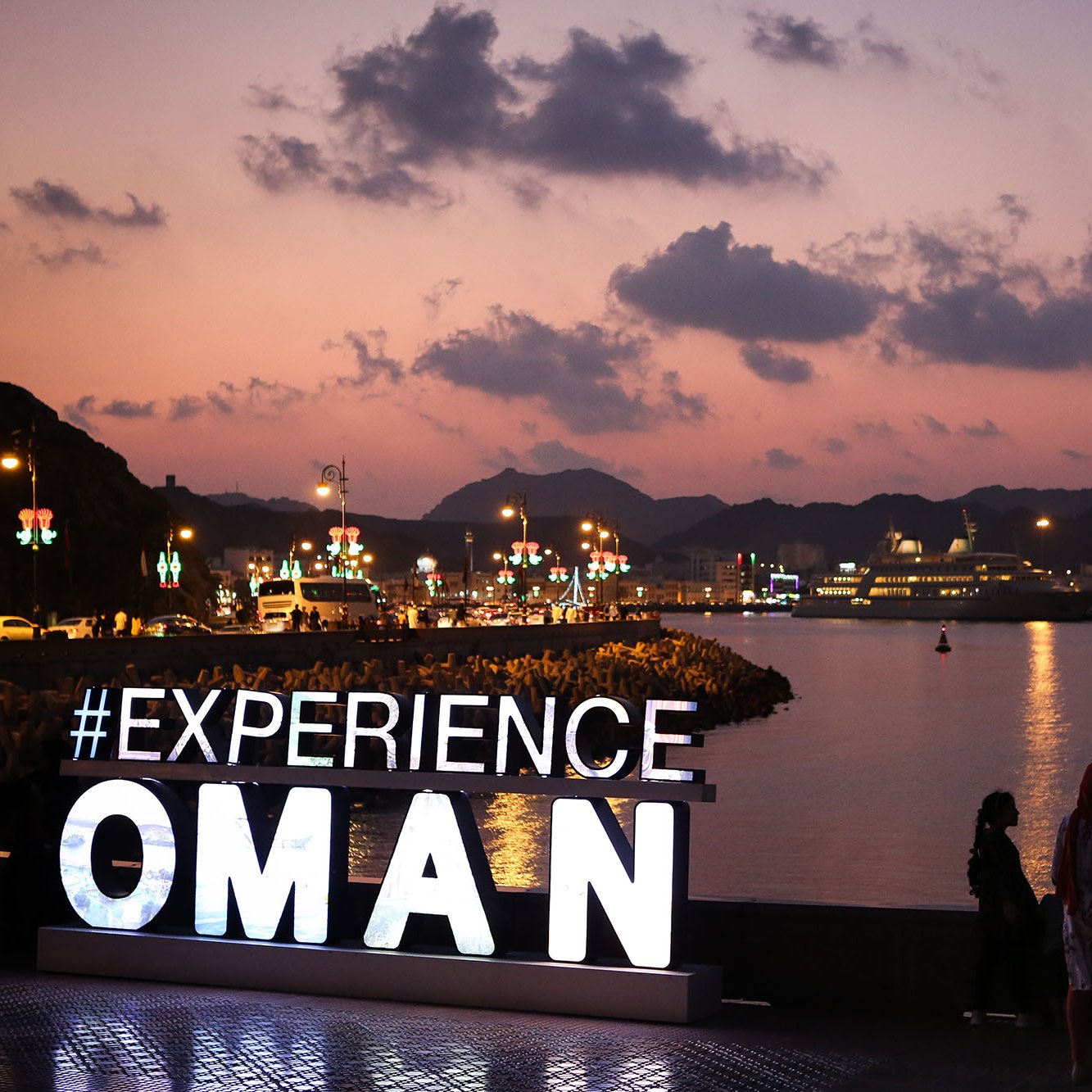 L'image montre une scène au coucher du soleil à Oman, avec des montagnes en arrière-plan et un port où se trouvent des bateaux et des navires de croisière. Au premier plan, il y a un grand panneau lumineux affichant le mot "EXPERIENCE" suivi d'"OMAN". Des personnes se tiennent devant le panneau, admirant la vue sur l'eau. La lumière douce du crépuscule et l'ambiance paisible de la scène créent une atmosphère accueillante.