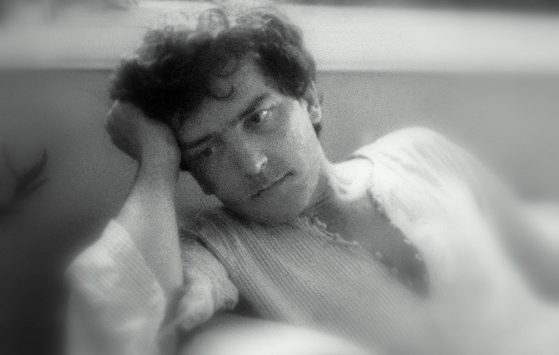 L'image montre un homme pensif, assis dans un endroit confortable. Il a les cheveux bouclés et un air réfléchissant, avec une main soutenant sa tête. Son expression semble mélancolique ou pensive. L'éclairage doux donne une ambiance nostalgique et calme à la scène. L'arrière-plan flou contribue à centrer l'attention sur son visage.