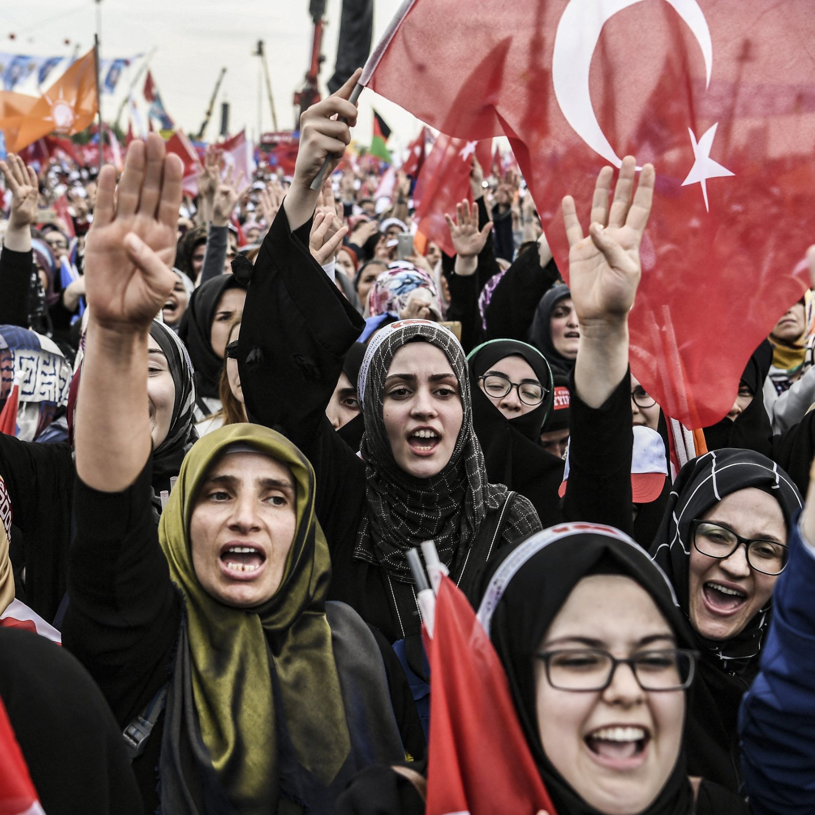 L'image montre une grande foule de personnes, principalement des femmes, qui sont rassemblées lors d'un événement ou d'une manifestation. Elles expriment leur enthousiasme et leur soutien en levant des drapeaux, dont certains arborent le symbole de la Turquie. Les participantes portent des vêtements traditionnels ou des foulards, et certaines semblent chanter ou crier des slogans. L'ambiance est dynamique et remplie d'énergie collective, suggérant une atmosphère de célébration ou de revendication.