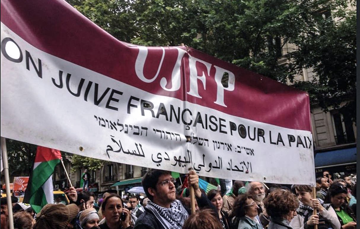 الصورة تظهر تجمعًا حاشدًا للأشخاص في مسيرة، حيث يرفع المشاركون لافتة كبيرة. اللافتة مكتوبة باللغات الفرنسية والعبرية والعربية، وتعبّر عن التضامن من أجل السلام. يبدو أن هناك تنوعًا في المشاركين، حيث يرتدي البعض الزي التقليدي أو يحملون أعلامًا مختلفة. الأجواء في الصورة تعكس روح النضال والمطالبة بحقوق معينة.