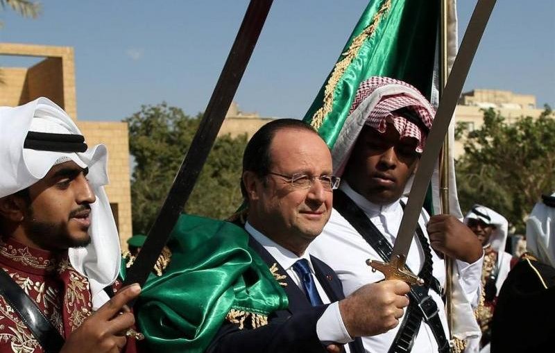 تظهر في الصورة مجموعة من الأشخاص يرتدون أزياء تقليدية. في المنتصف، يقف رجل يرتدي بدلة رسمية مع ابتسامة، محاطًا بشخصين آخرين يحمل كل منهما سيفًا. يزخر المشهد بالألوان، حيث يرتدي الشخصان الآخران ألبسة تقليدية بألوان بارزة، مع بعض الزينة الثقافية. الخلفية تضم أشجار وبعض المباني، وتبدو السماء صافية.