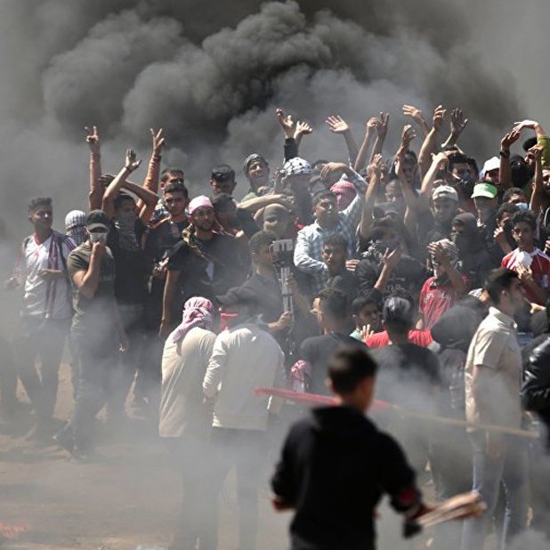 L'image montre un groupe de personnes rassemblées dans une atmosphère tendue. Elles sont entourées de fumée noire, probablement causée par des feux. Certains manifestants lèvent les bras en signe de protestation. On peut sentir une ambiance de colère et de résistance, avec quelques personnes qui semblent brandir des objets. Le décor est chaotique, évoquant une situation de crise ou de manifestation.