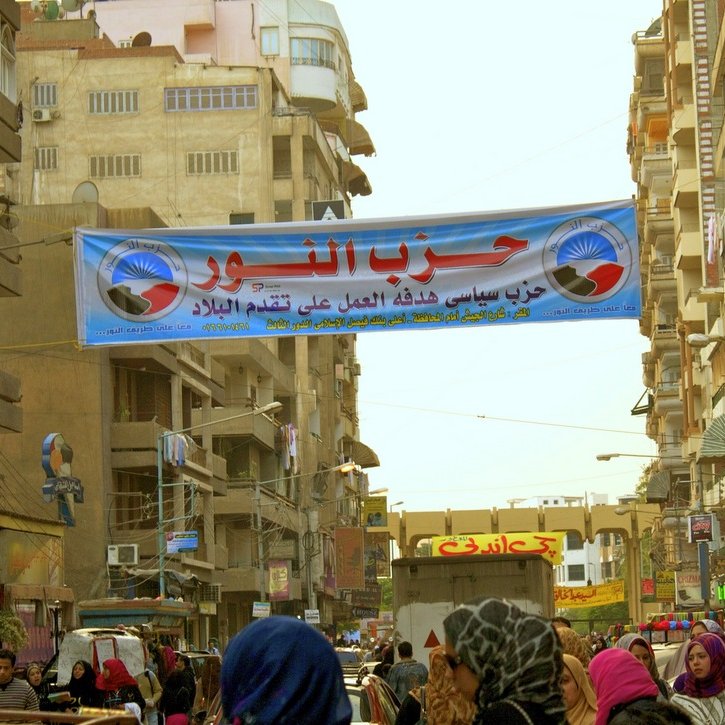 L'image montre une rue urbaine animée avec des bâtiments de plusieurs étages de chaque côté. Au-dessus de la route, il y a une grande banderole avec du texte en arabe. Cette banderole semble promouvoir un parti politique, qui pourrait être lié à des événements ou des activités locales. On aperçoit également des personnes marchant dans la rue, certaines portant des vêtements traditionnels. L'atmosphère de l'image évoque l'animation d'un marché ou d'un rassemblement communautaire.