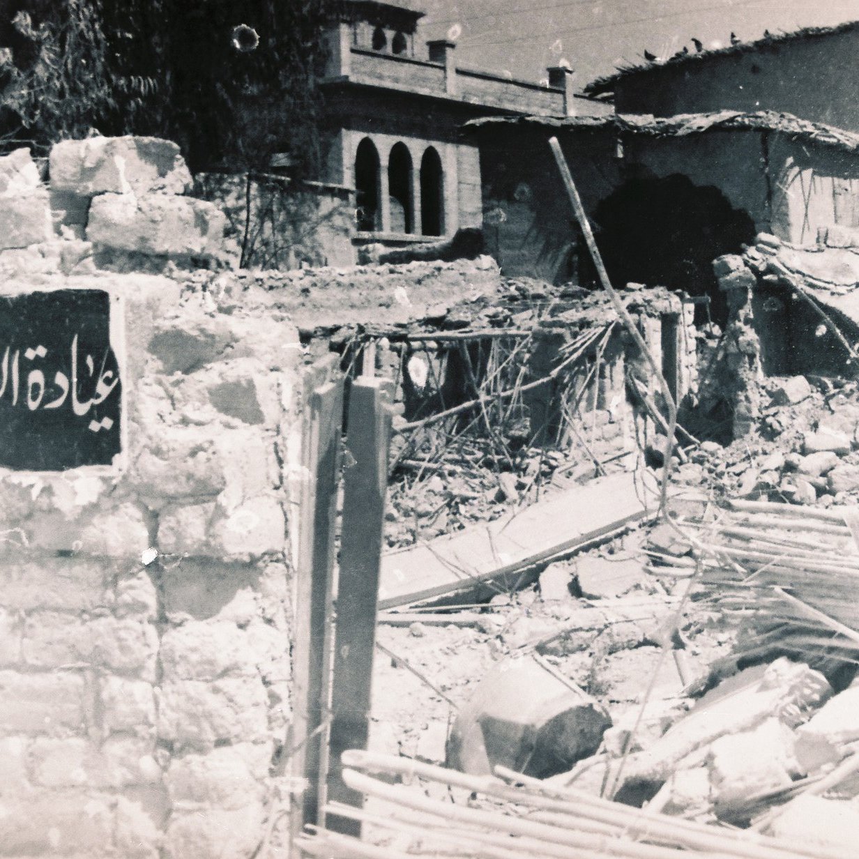 الصورة تُظهر مشهداً لمدمرات بسبب أحداث سابقة، حيث يظهر حطام المباني والخرائب. في مقدمة الصورة، توجد لافتة تحمل اسم مكان، ولكنها مهملة وسط الأنقاض. الخلفية تحتوي على نوافذ وأجزاء من مباني قديمة، مما يعكس تأثير الزمن أو كارثة معينة. الأجواء توحي بالحزن والألم الناتج عن الدمار.