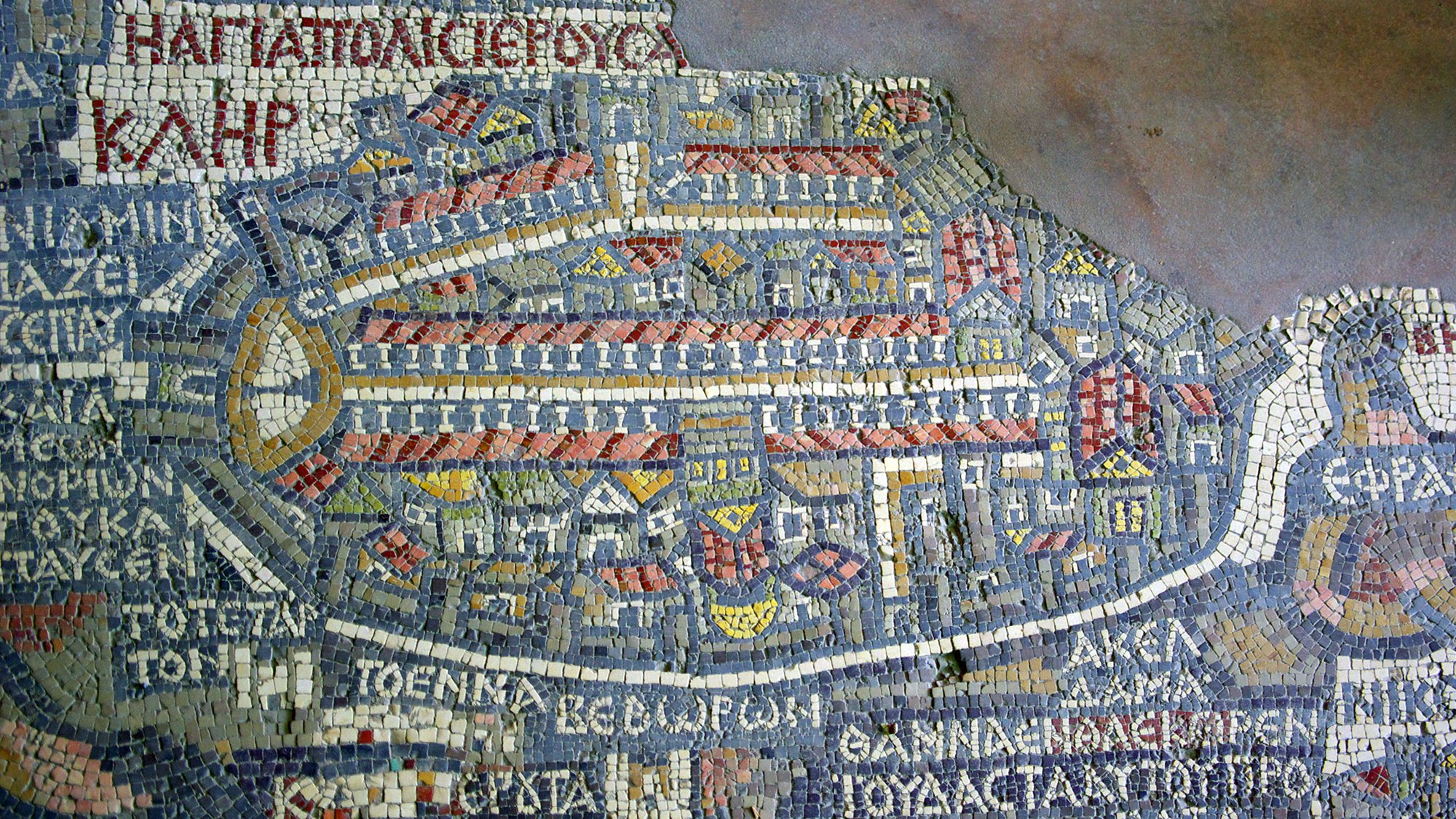 The image depicts a mosaic map, featuring intricate tile work that illustrates an ancient city layout. The map is composed of various colored tiles, forming buildings, streets, and outlines of the architecture within the city. It includes labeled regions or areas, indicating places of significance in the city, which might be historical or prominent during the time it represents. The overall design reflects a detailed representation of urban planning, showcasing the artistry and craftsmanship of the era.