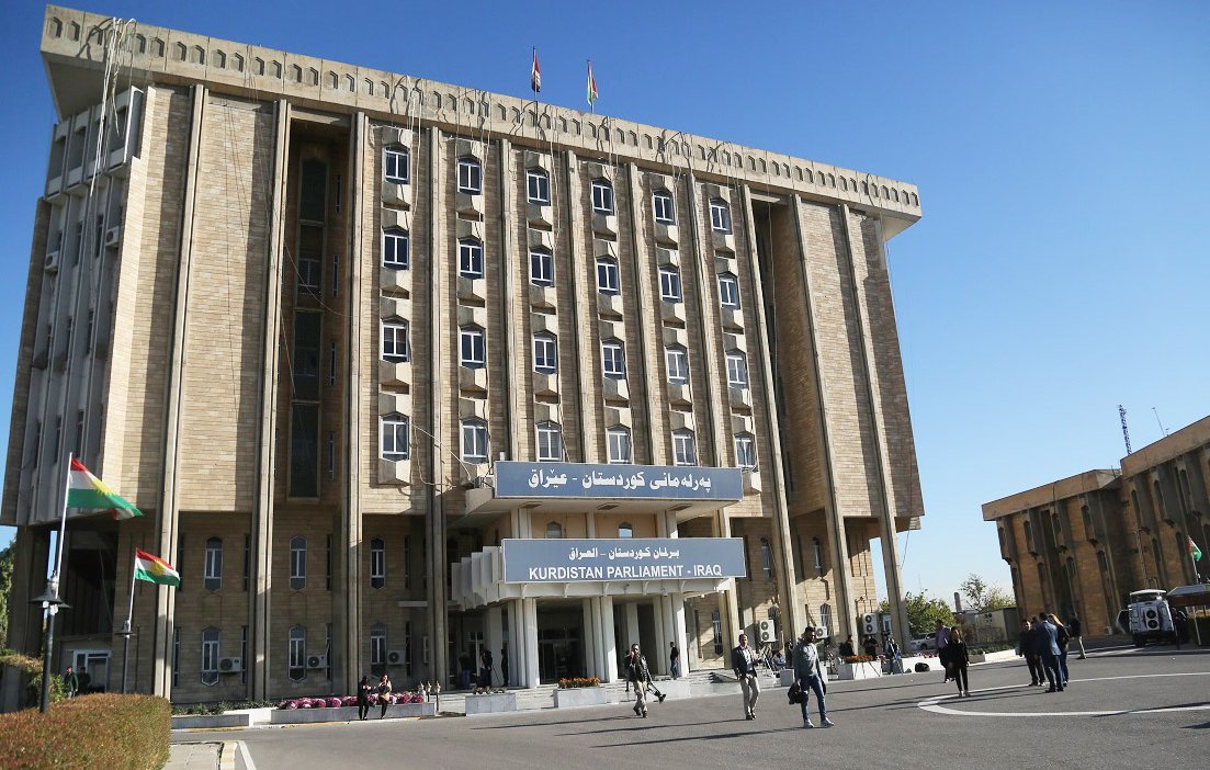 این تصویر نمای ساختمان پارلمان کردستان در عراق را نشان می‌دهد. ساختمان دارای طراحی مدرن و بزرگ است و چندین طبقه دارد. در جلوی ساختمان، پرچم‌های کردستان و عراق به اهتزاز درآمده‌اند. همچنین، افرادی در حال تردد در محوطه‌ مقابل ساختمان دیده می‌شوند. آسمان آبی و صاف به زیبایی محیط افزوده است.