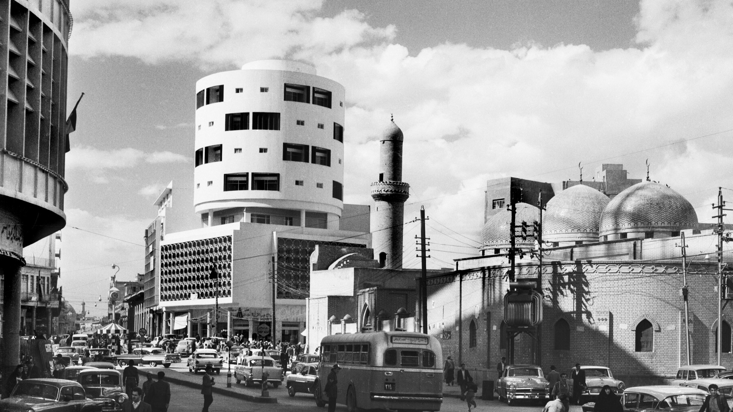 L'image en noir et blanc présente une scène urbaine animée. Au premier plan, on peut voir une rue avec plusieurs véhicules, dont des voitures et un bus. Le bâtiment à gauche a une architecture moderne, tandis qu'à droite, on aperçoit un édifice avec des dômes caractéristiques, suggérant une influence architecturale traditionnelle. Des piétons circulent, et on remarque également un homme sur un vélo. Le ciel est partiellement nuageux, ajoutant à l'atmosphère de la scène. Cette image évoque un mélange d'architecture moderne et traditionnelle dans un contexte urbain.