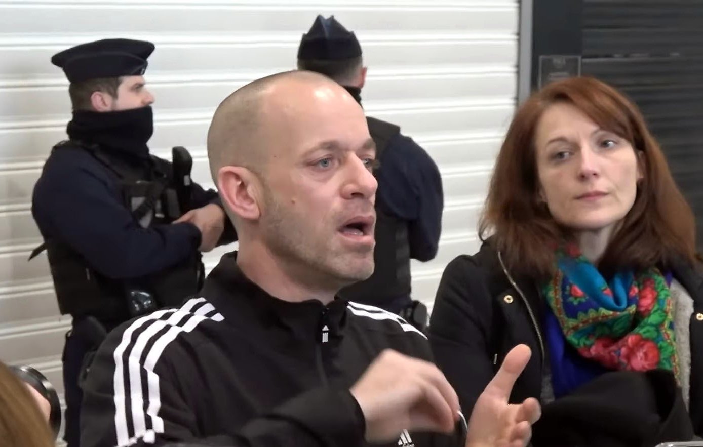 L'image montre un homme qui parle, visiblement en train d'expliquer quelque chose avec des gestes. Il porte un survêtement noir avec des rayures blanches. À côté de lui, une femme l'écoute attentivement, les cheveux bruns et portant un manteau noir. À l'arrière-plan, on peut voir des policiers en uniforme, certains portant des masques. L'ensemble suggère une situation de discussion ou d'intervention, possiblement liée à un événement impliquant les forces de l'ordre.