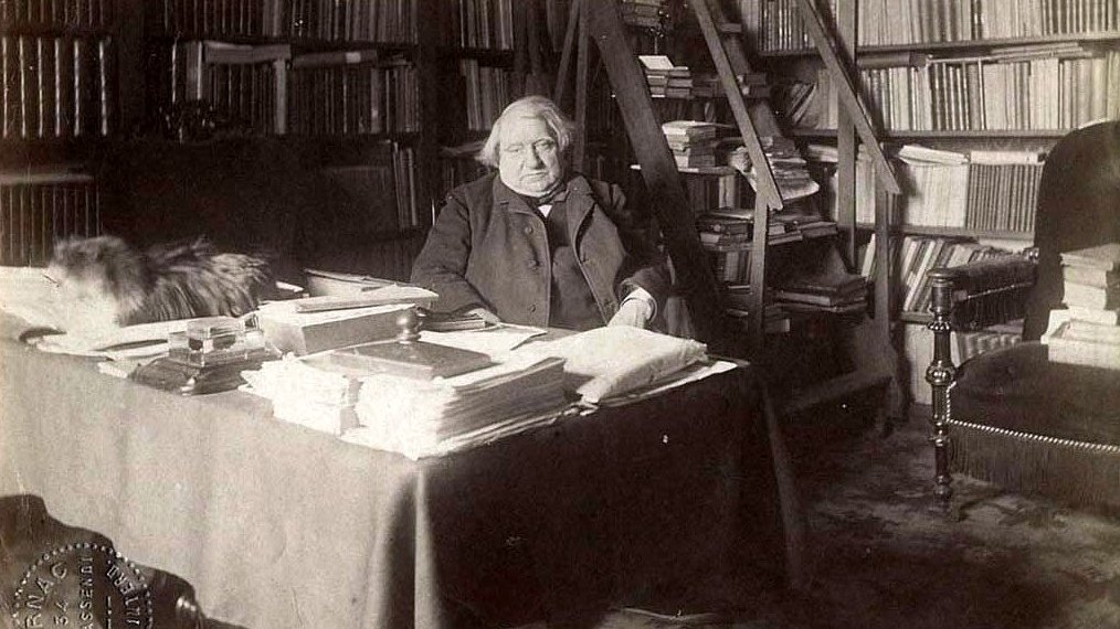 L'image montre une scène d'intérieur, probablement dans un bureau ou une bibliothèque. Un homme, apparemment âgé, est assis à une table chargée de papiers et de livres. Derrière lui, on peut voir des étagères remplies de livres. Il y a également un tapis sur le sol. Un chat, qui semble être sur la table ou à proximité, ajoute une touche de vie à l'atmosphère studieuse de la pièce. L'éclairage donne une impression de sérénité et de calme.