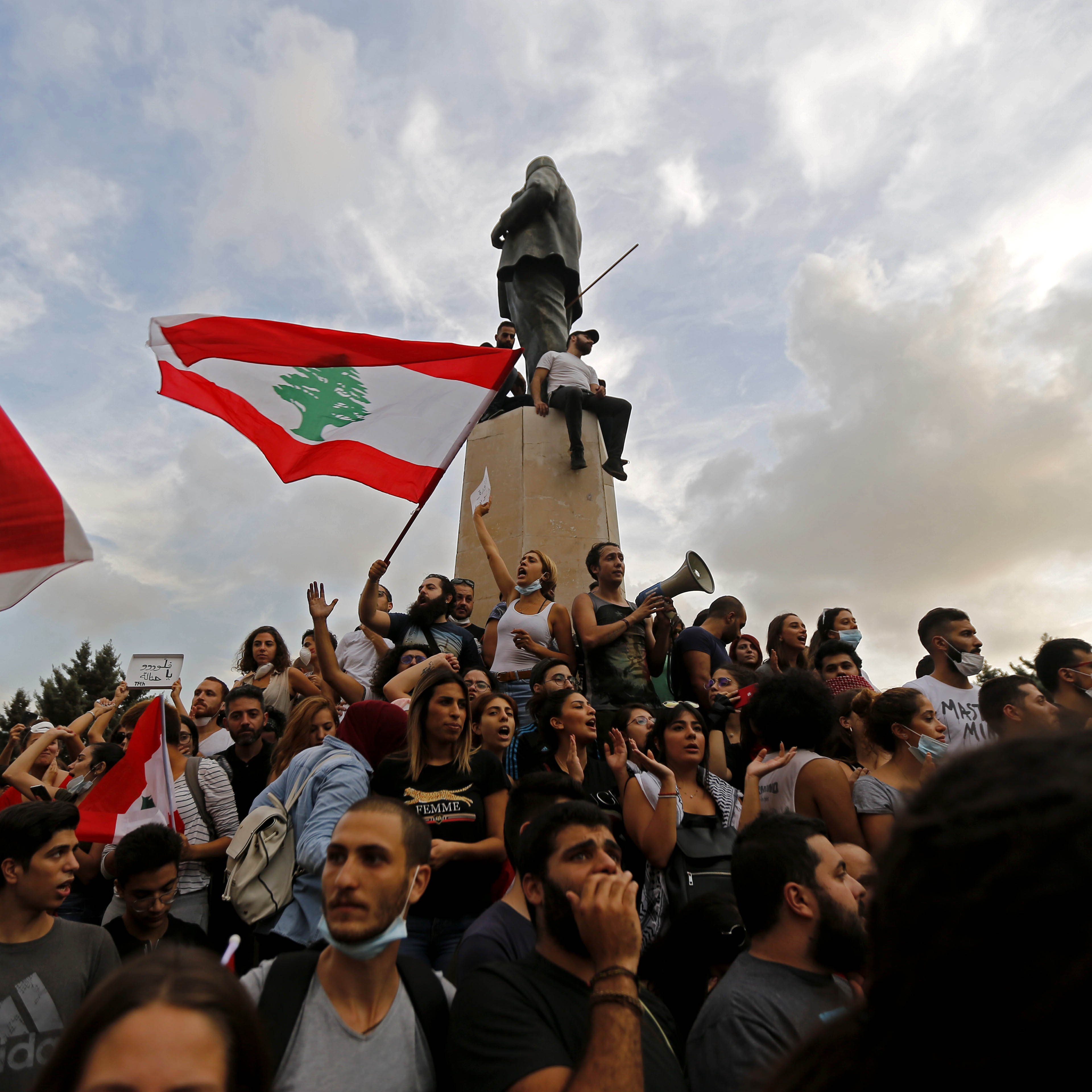 L'image montre une manifestation au Liban. Des groupes de personnes sont rassemblés autour d'un monument, tenant des drapeaux libanais. Certains manifestants chantent ou s'expriment à l'aide de mégaphones. L'atmosphère semble dynamique et engagée, avec des nuages dans le ciel qui ajoutent une ambiance dramatique à la scène.