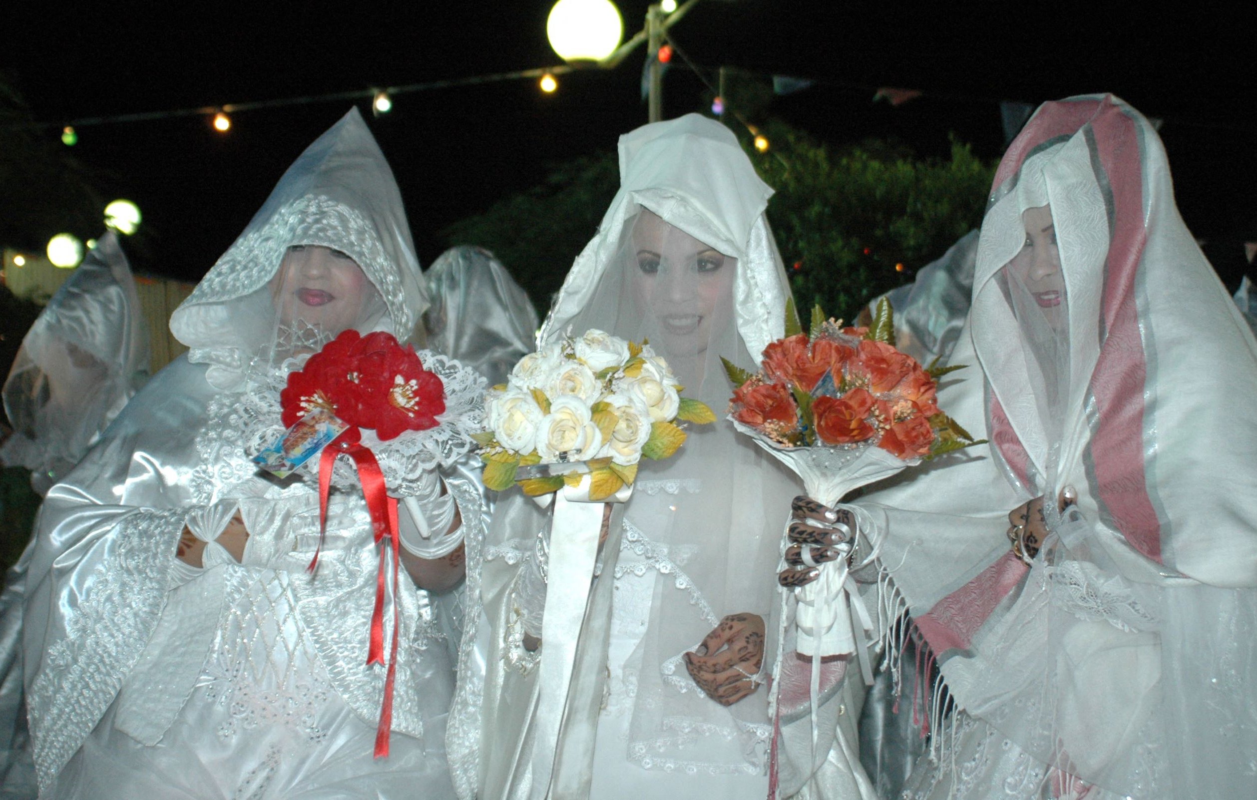 L'image montre un groupe de femmes vêtues de robes blanches et de voiles, se tenant ensemble dans une ambiance nocturne. Elles tiennent chacune des bouquets de fleurs, composés de roses et d'autres fleurs colorées. L'éclairage doux crée une atmosphère festive, et des guirlandes lumineuses sont visibles en arrière-plan. Leurs visages sont en partie cachés par les voiles, ajoutant une aura de mystère à la scène.