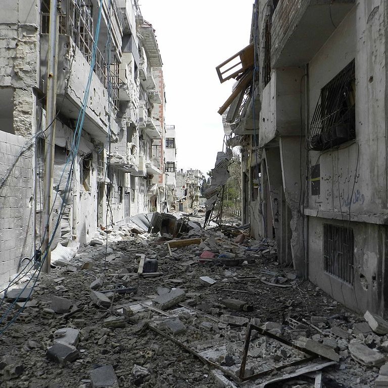L'image montre une rue dévastée, probablement dans une zone de conflit. Les bâtiments de chaque côté sont endommagés, avec des murs effondrés et des débris éparpillés sur le sol. Des fils électriques pendent, et l'atmosphère générale dégage une impression de destruction et d'abandon. On peut observer des traces des conséquences des affrontements, avec un paysage urbain sévèrement altéré.