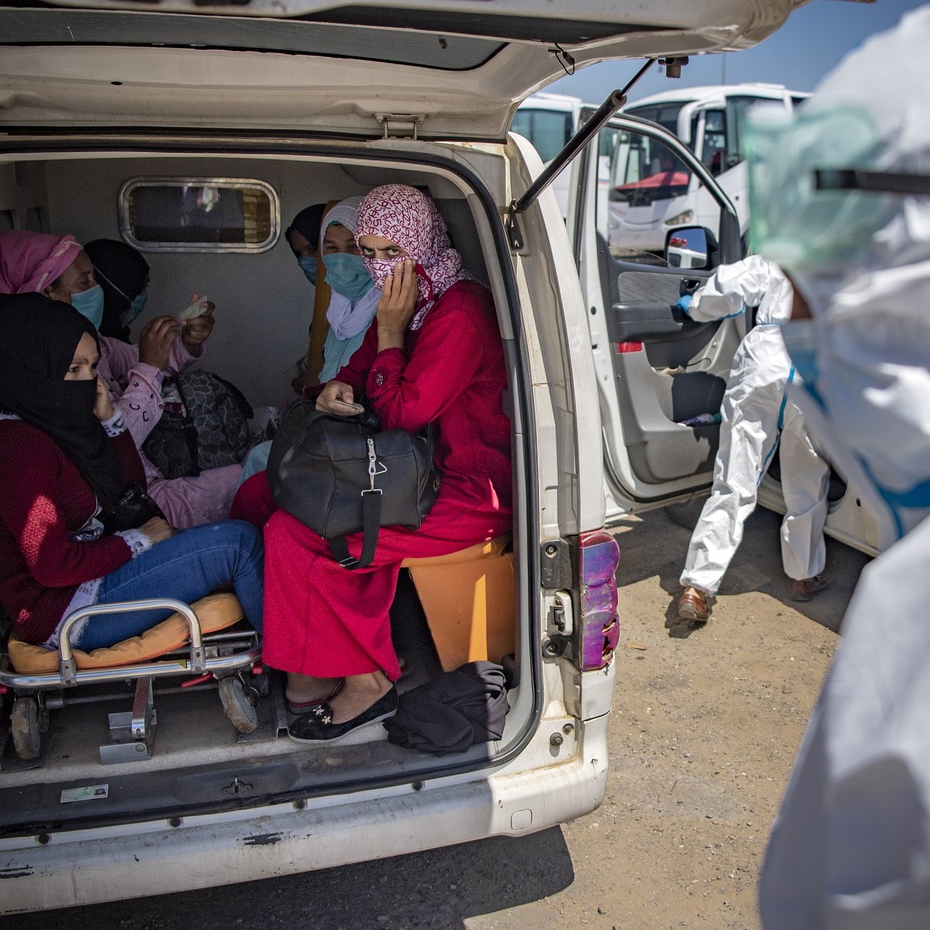 L'image montre un groupe de personnes à l'intérieur d'une ambulance. On peut voir plusieurs femmes assises, certaines portant des masques et des vêtements colorés. Une femme porte un hijab et semble discuter avec les autres. À l'extérieur, une personne en tenue de protection, probablement un professionnel de la santé, s'approche. L'environnement semble être une zone d'intervention médicale. La scène évoque une atmosphère de soin et d'assistance dans un contexte de santé.
