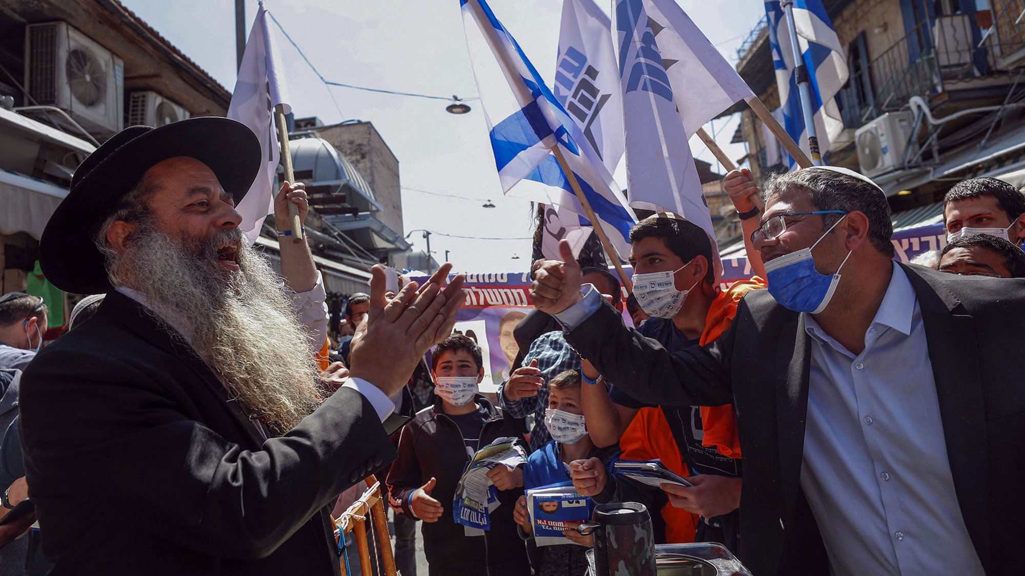 L'image montre une scène animée dans une rue, probablement lors d'un rassemblement ou d'une manifestation. Des personnes se tiennent debout, brandissant des drapeaux, apparemment en soutien à une cause. Au centre, un homme avec une longue barbe et un chapeau noir s'adresse à un autre homme, qui semble engagé dans un échange passionné. Les deux semblent entourés par une foule de jeunes gens, certains portant des masques de protection. L'environnement urbain et les bâtiments en arrière-plan ajoutent à l'atmosphère dynamique de l'événement.