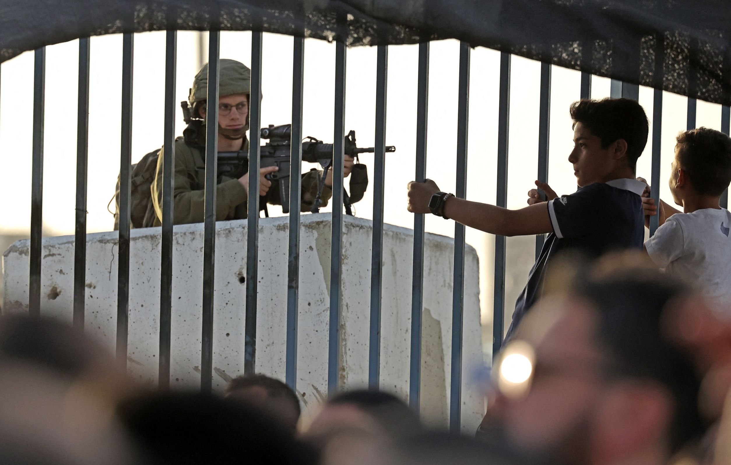 L'image montre un soldat armé stationné derrière une barrière ou un grillage. Il est en position de vigilance, regardant au loin. En face de lui, un jeune garçon semble gesticuler, tandis que d'autres enfants ou adolescents se trouvent à proximité. L'environnement semble tendu, reflétant une situation de contrôle ou de conflit. La lumière du soleil indique un moment de la journée où l'activité est encore présente.