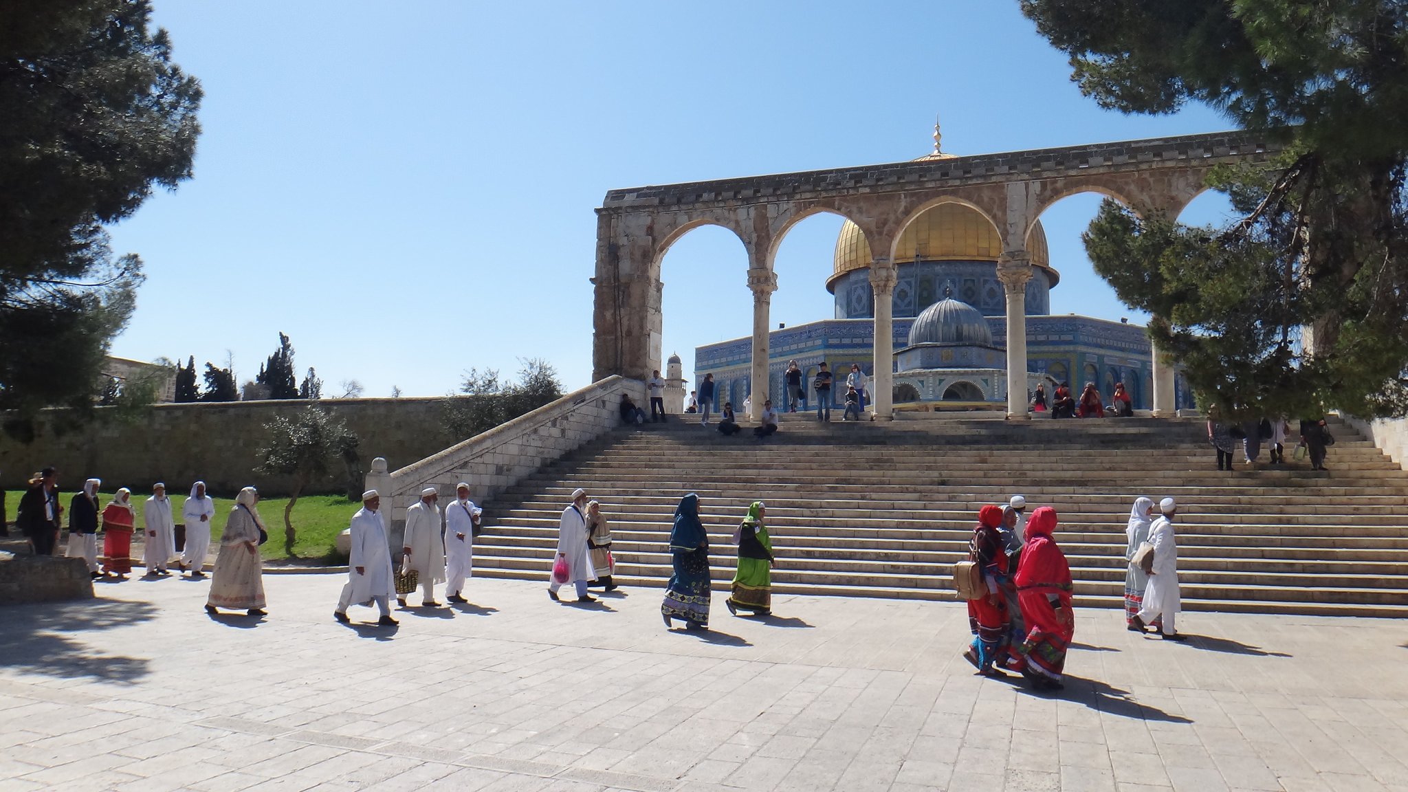 L'image montre un paysage architectural impressionnant avec des arches majestueuses en premier plan. On peut voir un groupe de personnes marchant sur une large marche, certaines portant des vêtements traditionnels colorés. En arrière-plan, la célèbre mosquée avec son dôme doré est visible, entourée d'un ciel clair et d'arbres verdoyants. L'atmosphère semble paisible et spirituelle, reflet d'un lieu de rassemblement important.