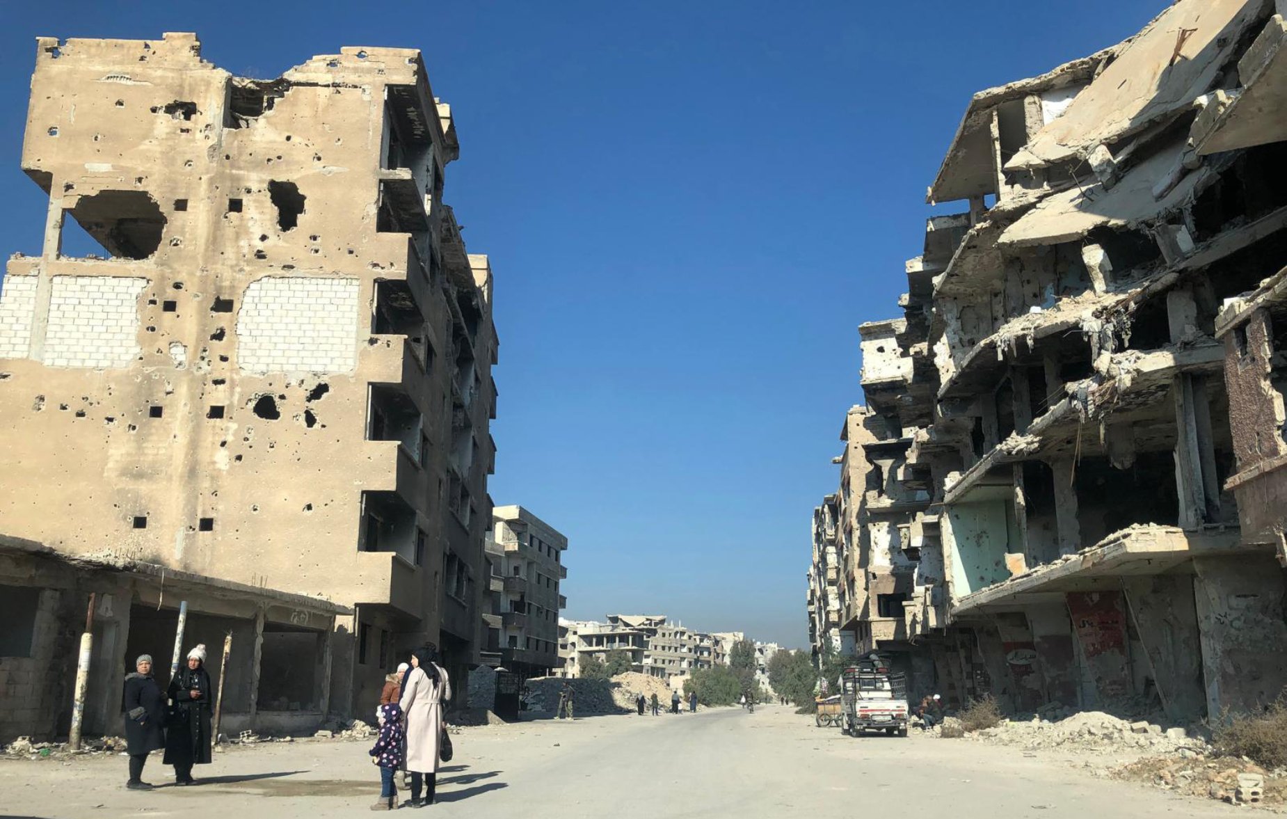 تصویر یک خیابان خالی را نشان می‌دهد که در دو طرف آن ساختمان‌های مخروبه و آسیب‌دیده قرار دارند. دیوارهای این ساختمان‌ها پر از سوراخ‌ها و آسیب‌های ناشی از جنگ یا تخریب هستند. در انتهای خیابان، چند ساختمان دیگر نیز مشاهده می‌شود. آسمان آبی و صاف است و افراد کمی در خیابان در حال تردد هستند. دو نفر در سمت چپ تصویر به نظر می‌رسد در حال صحبت کردن هستند و یک کودک نیز با آن‌هاست. فضای کلی تصویر تنهایی و تخریب را القا می‌کند.