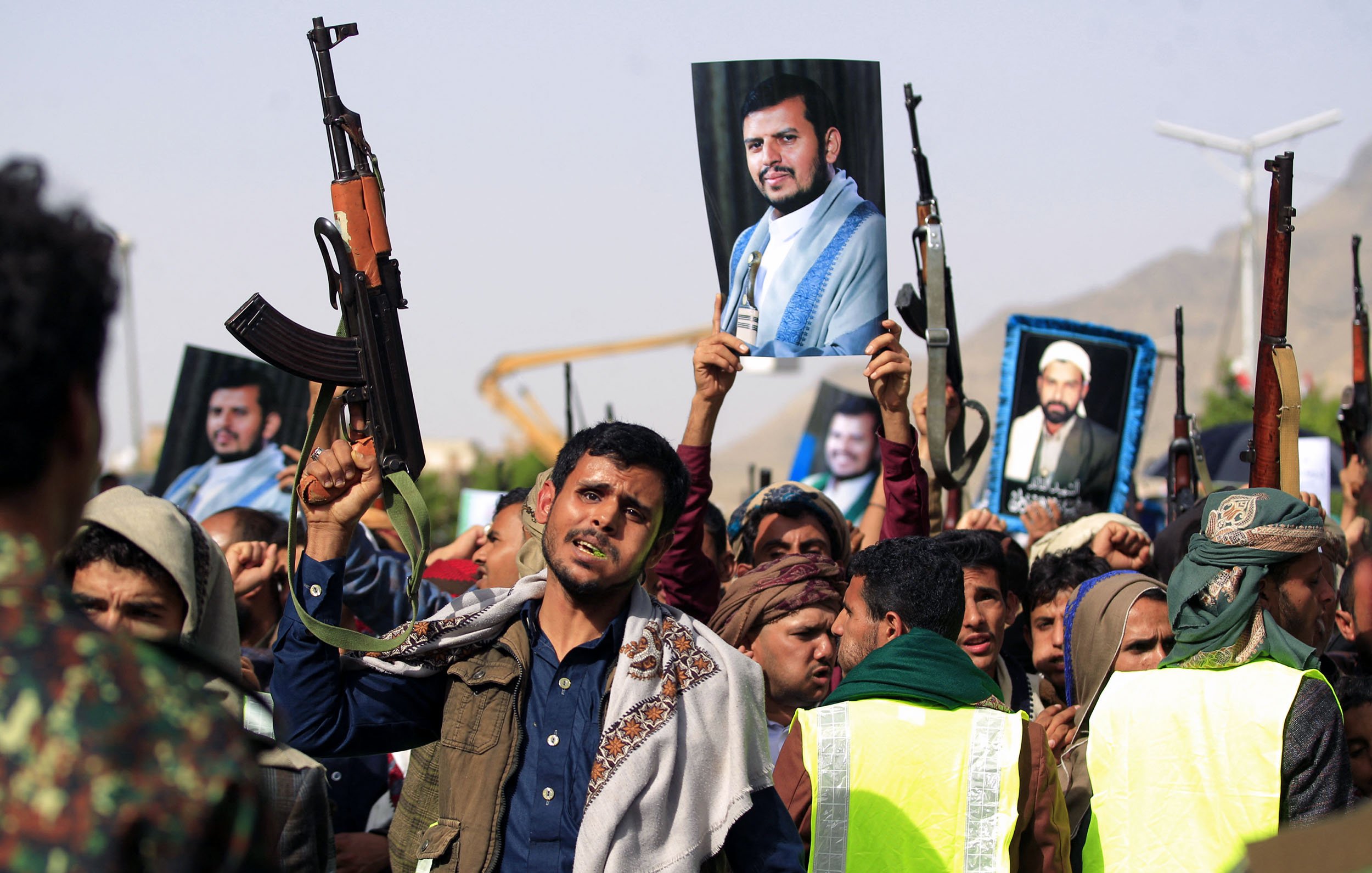L'image montre une foule de manifestants tenant des fusils automatiques. Certains d'entre eux brandissent des portraits de figures emblématiques. Les gens semblent exprimer des émotions fortes, comme la détermination ou l'enthousiasme. L'environnement évoque une ambiance de rassemblement ou de manifestation politique. Des éléments comme des vêtements spécifiques et le paysage suggèrent un contexte culturel particulier.