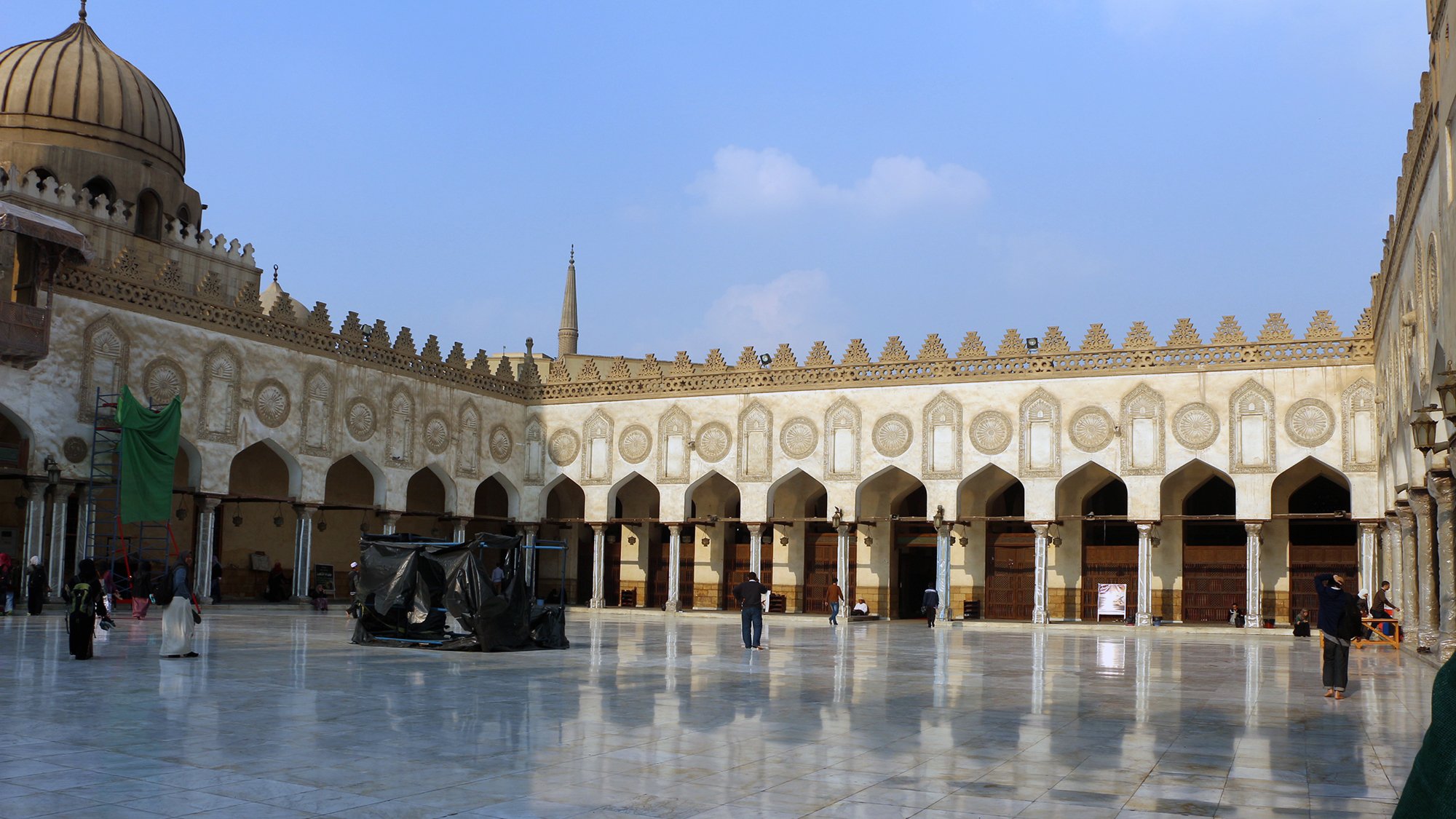 L'image montre une cour spacieuse d'un bâtiment historique, probablement un lieu de culte ou une mosquée. Le sol est en marbre poli, reflétant la lumière du ciel. On peut observer des arches élégantes et de nombreuses colonnes tout autour, ornées de motifs architecturaux détaillés. Au fond, on aperçoit un dôme et des minarets qui s'élèvent dans le ciel. La scène est calme, avec quelques personnes se déplaçant dans l'espace. Le ciel est clair, parsemé de nuages légers.