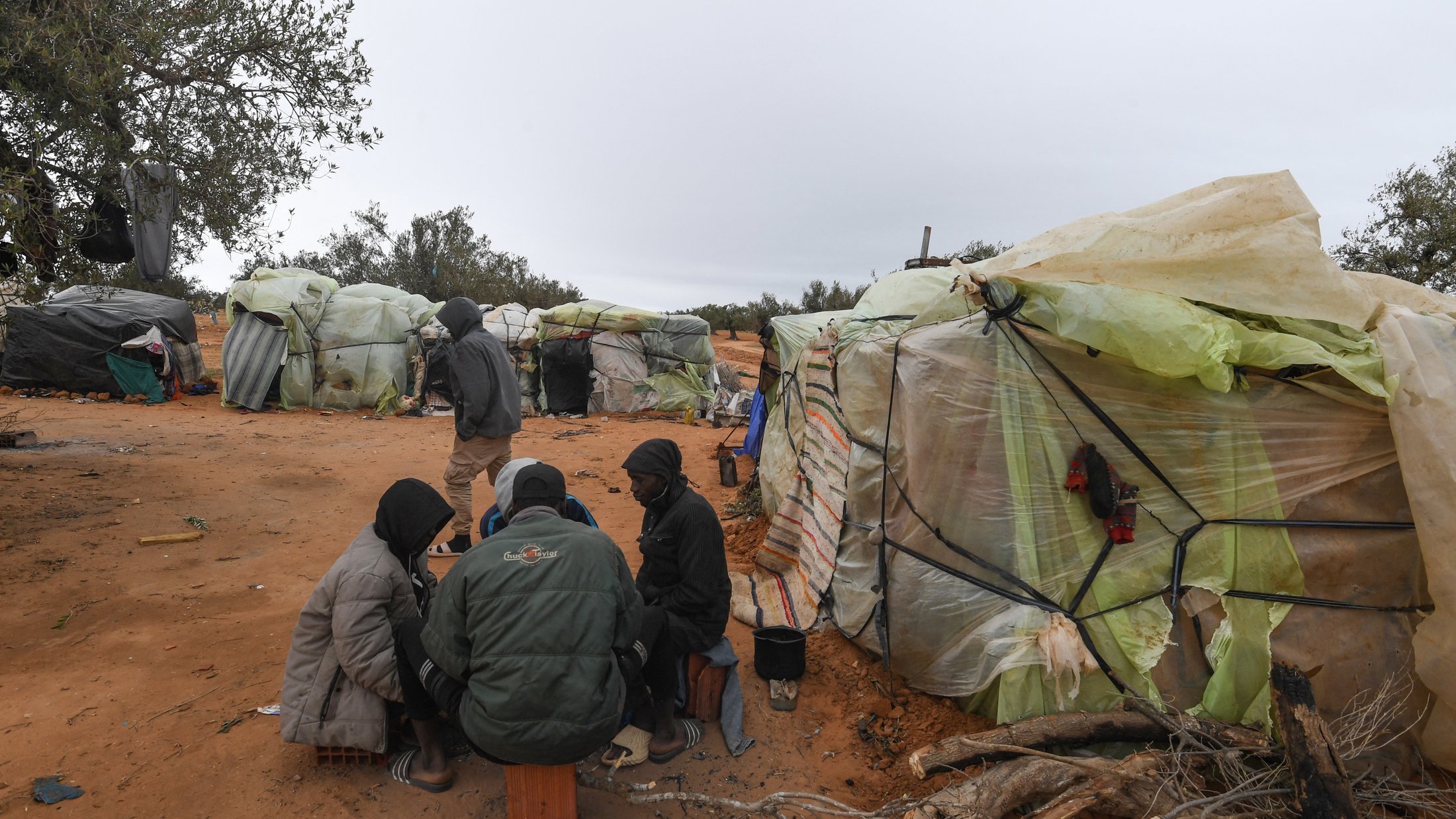 L'image montre un groupe de personnes assises ensemble sur le sol, entourées de tentes faites de matériaux recyclés, telles que des bâches en plastique. Le décor est désertique, avec un sol de sable et quelques arbres à l'arrière-plan. Les personnes semblent être en conversation ou en train de partager quelque chose. Le temps paraît nuageux, ajoutant une atmosphère sombre à la scène.