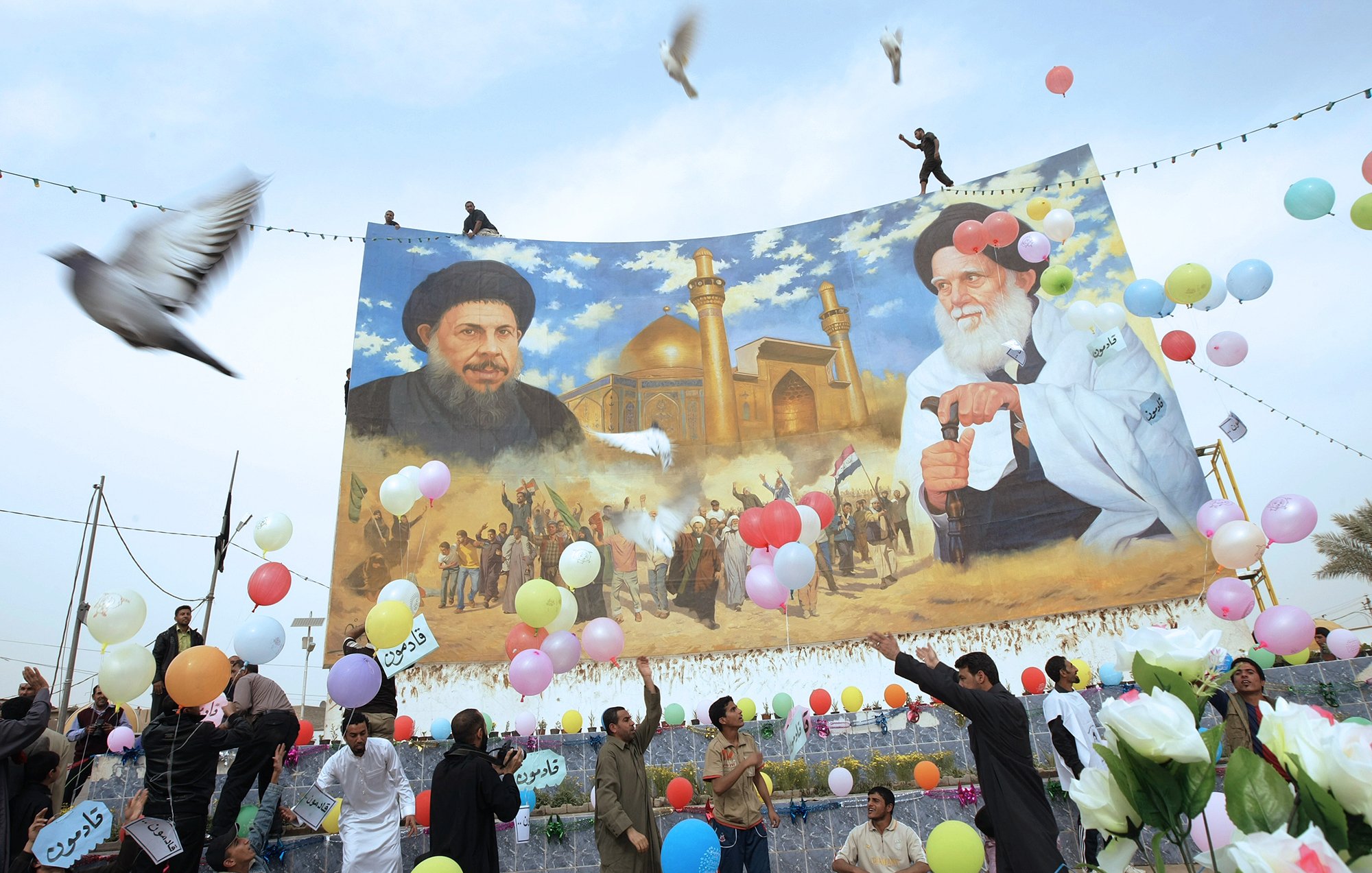 در این تصویر، تعدادی از مردم در حال برگزاری یک مراسم یا جشن هستند. در پس‌زمینه، یک دیوار بزرگ با تصویری از دو شخصیت سیاسی یا مذهبی به نمایش درآمده است. افراد در حال پرتاب بالن‌های رنگی و همچنین برخی گل‌ها به سمت هوا هستند. برخی از مردم در حال رقص و شادمانی دیده می‌شوند. پرنده‌هایی نیز در آسمان در حال پرواز هستند و فضای کلی تصویر روحیه مثبت و شادابی را به نمایش می‌گذارد.