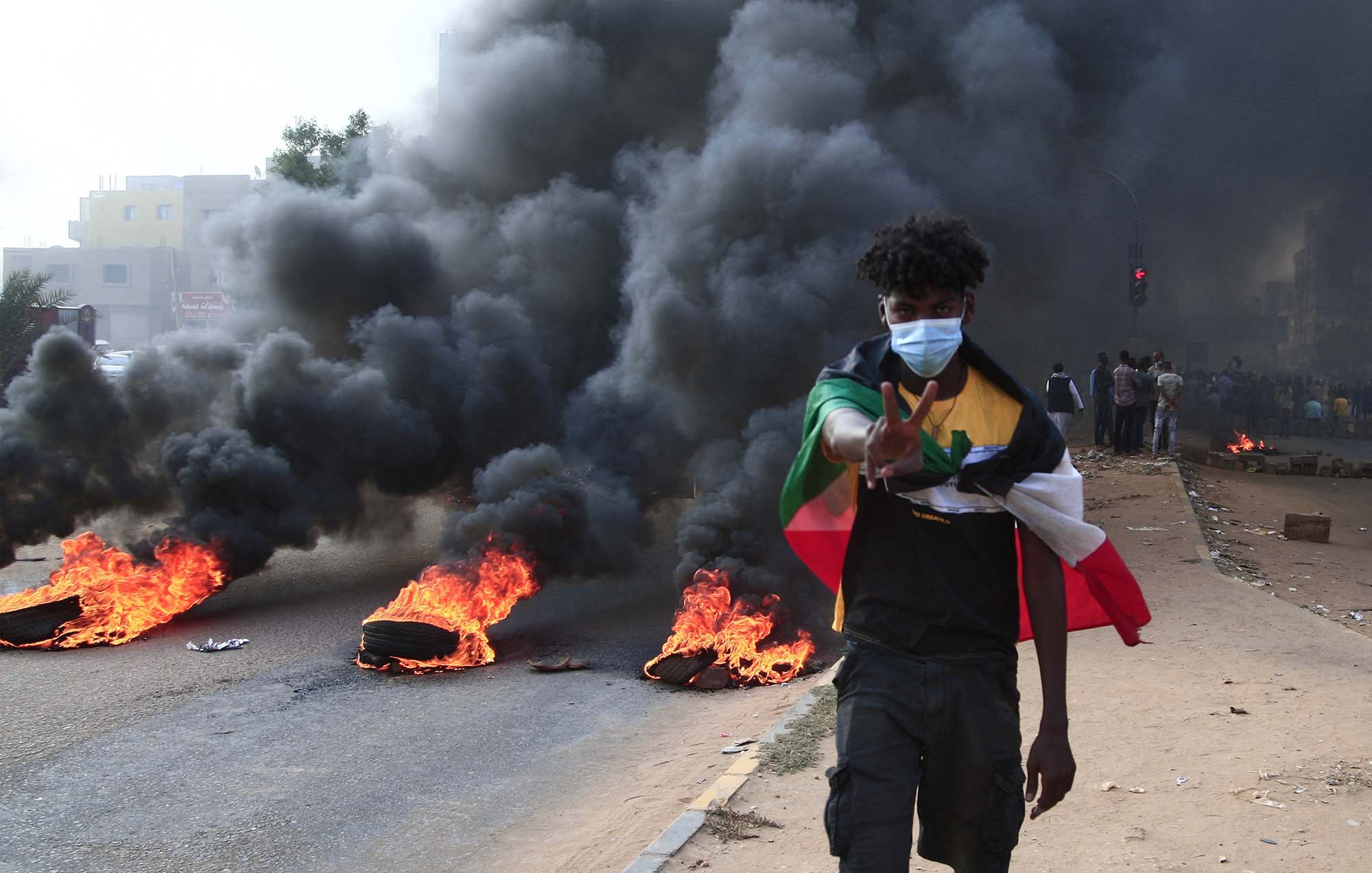 L'immagine mostra un giovane che cammina lungo una strada, con fumi neri che si alzano dietro di lui. Ci sono pneumatici in fiamme che bruciano ai lati della strada. Il ragazzo indossa una mascherina e fa un gesto di pace con la mano. Sullo sfondo, si notano altre persone che osservano la scena. L'atmosfera sembra intensa e carica di tensione.