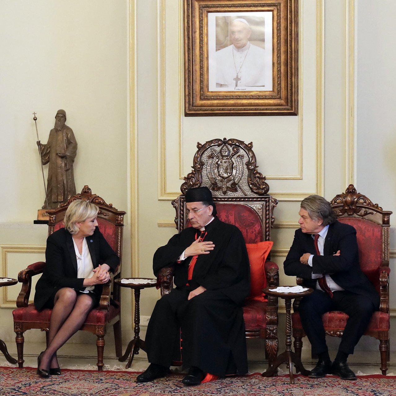 L'image montre une scène d'une réunion ou d'un entretien formel. Trois personnes sont assises dans des fauteuils ornés, au milieu d'une pièce décorée de manière élégante. L'une des personnes, portant une tenue ecclésiastique, semble être en train de parler, tandis que les autres l'écoutent attentivement. Les murs sont ornés de statues et d'œuvres d'art, ce qui suggère un environnement culturel et riche en histoire.