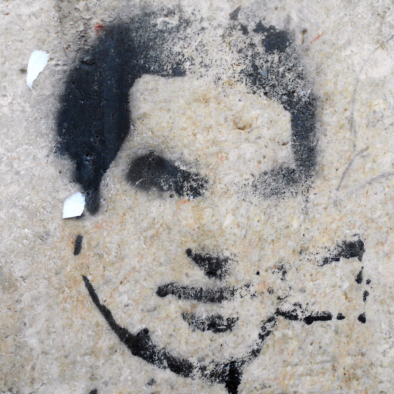 L'image montre un pochoir sur un mur, représentant un visage stylisé. Les contours sont en noir, avec un fond de pierre claire. Le visage semble être celui d'une personne jeune, avec une expression neutre. Quelques résidus de papier sont éparpillés autour, ajoutant une texture supplémentaire au mur. L'ensemble dégage une atmosphère urbaine et artistique.