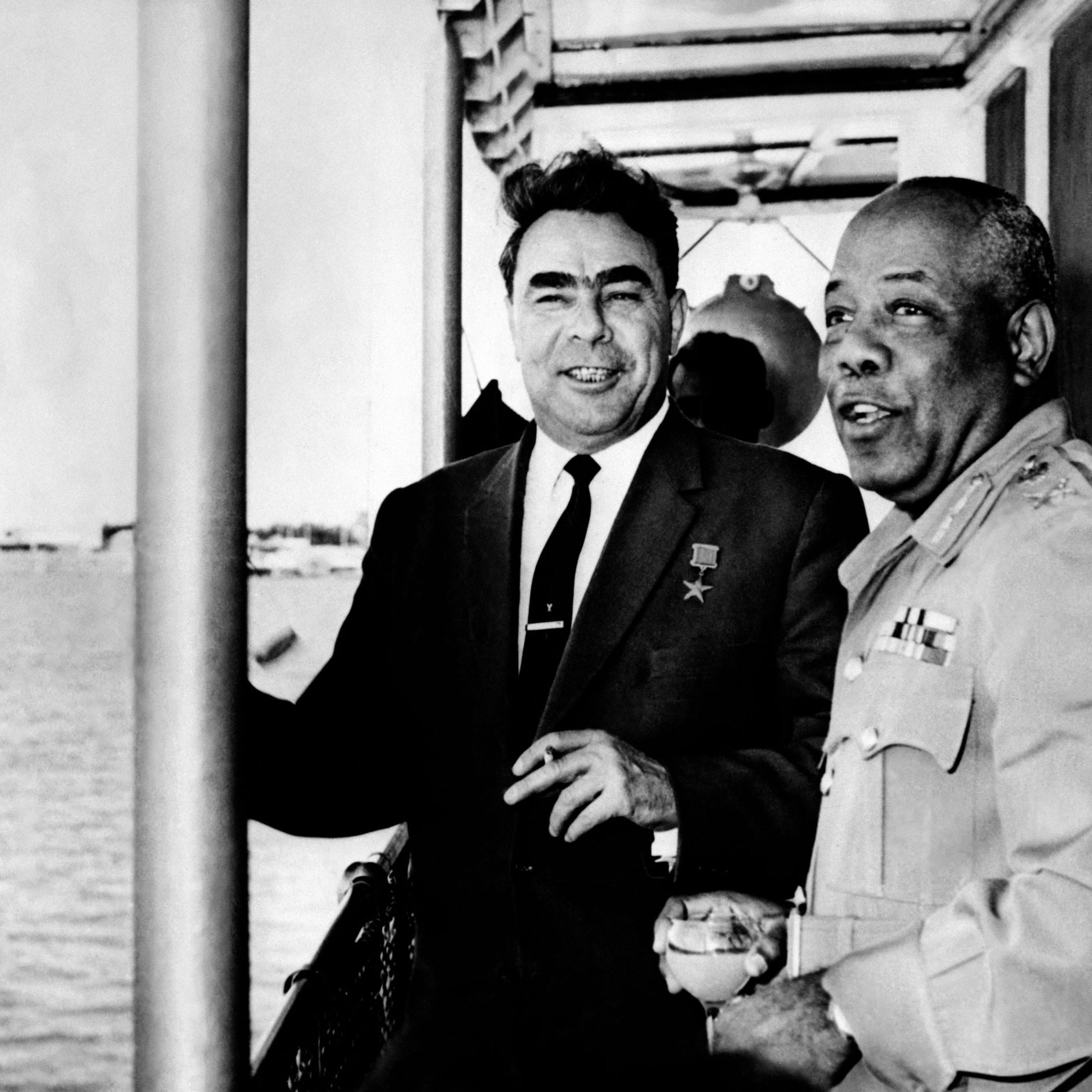 L'image montre deux hommes en train de discuter sur ce qui semble être un bateau ou une plateforme près de l'eau. L'un des hommes porte une médaille, indiquant honneur ou reconnaissance, et les deux semblent souriants et détendus. En arrière-plan, on peut voir des bateaux et une vue maritime. L'atmosphère générale de l'image semble amicale et conviviale.