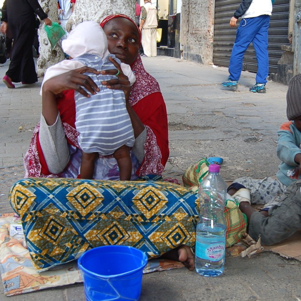 L'image montre une femme assise sur le sol, tenant un bébé dans ses bras. Elle est entourée de deux enfants, l'un assis à côté d'elle et l'autre jouant sur le sol. La femme porte un vêtement coloré et un foulard, tandis que les enfants semblent habillés de manière simple et modeste. À côté d'eux, il y a une bouteille d'eau et un seau bleu. La scène se déroule dans une rue, avec des bâtiments en arrière-plan et d'autres personnes qui passent. L'ambiance semble à la fois familiale et difficile, évoquant des thèmes de pauvreté et de lutte quotidienne.