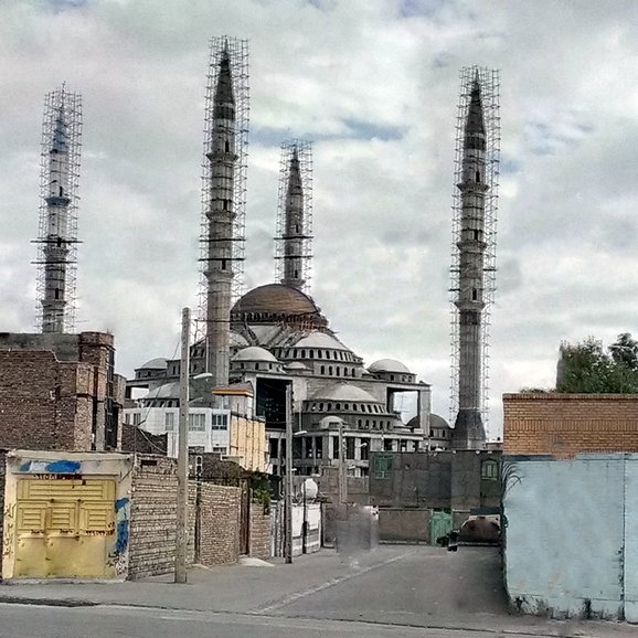 L'image montre un grand bâtiment au centre, semblable à une mosquée, avec plusieurs minarets élancés qui s'élèvent vers le ciel. Le bâtiment a un dôme proéminent et semble être en construction ou en rénovation, avec des échafaudages visibles. En arrière-plan, le ciel est nuageux, ce qui donne à la scène une atmosphère un peu sombre. À l'avant, on peut apercevoir des structures et des murs en briques, ainsi qu'une route. L'ensemble de la composition évoque un mélange d'architecture traditionnelle et moderne.