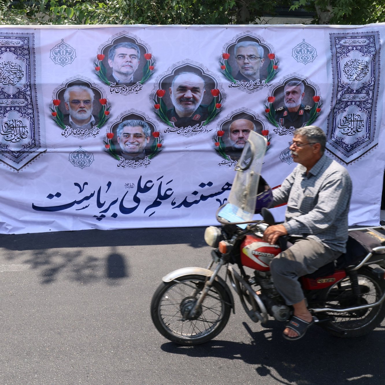 تصویر مردی سوار بر موتورسیکلت که در پیش‌زمینه، بنر بزرگ و مردم نشسته در پشت آن دیده می‌شوند.